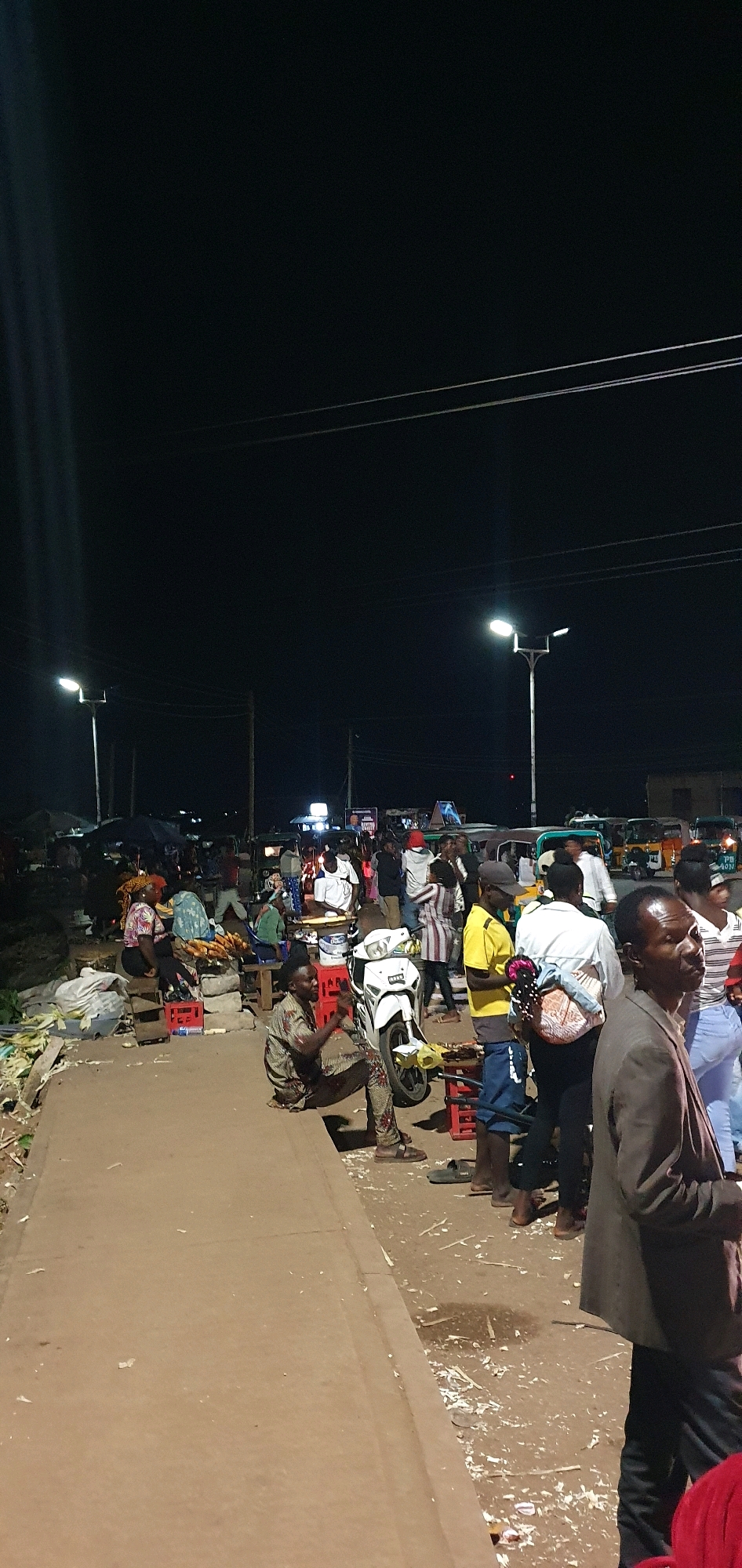 The image depicts a bustling night market scene in Bukuru, Nigeria. The foreground is dominated by a paved walkway with several people standing and sitting. A man in a patterned shirt and pants is sitting on the ground near a white motorcycle, appearing to be interacting with it. To his right, another man in a brown jacket is looking away from the camera, and a woman in a striped top is walking past.
Further into the scene, several market stalls are set up, with people gathered around them. Vendors appear to be selling goods, with some items, possibly food like corn on the cob, visible on display. There are several red crates scattered around, some used as seating or to elevate goods. A white motorcycle is parked centrally in the mid-ground.
The background reveals a busy street with multiple vehicles, including what appear to be tricycles or tuk-tuks, suggesting active transportation. Streetlights illuminate the area, casting light on the crowds and stalls. The overall atmosphere is lively and typical of a nocturnal marketplace in a Nigerian city. The environment appears to be an open-air market on a roadside.