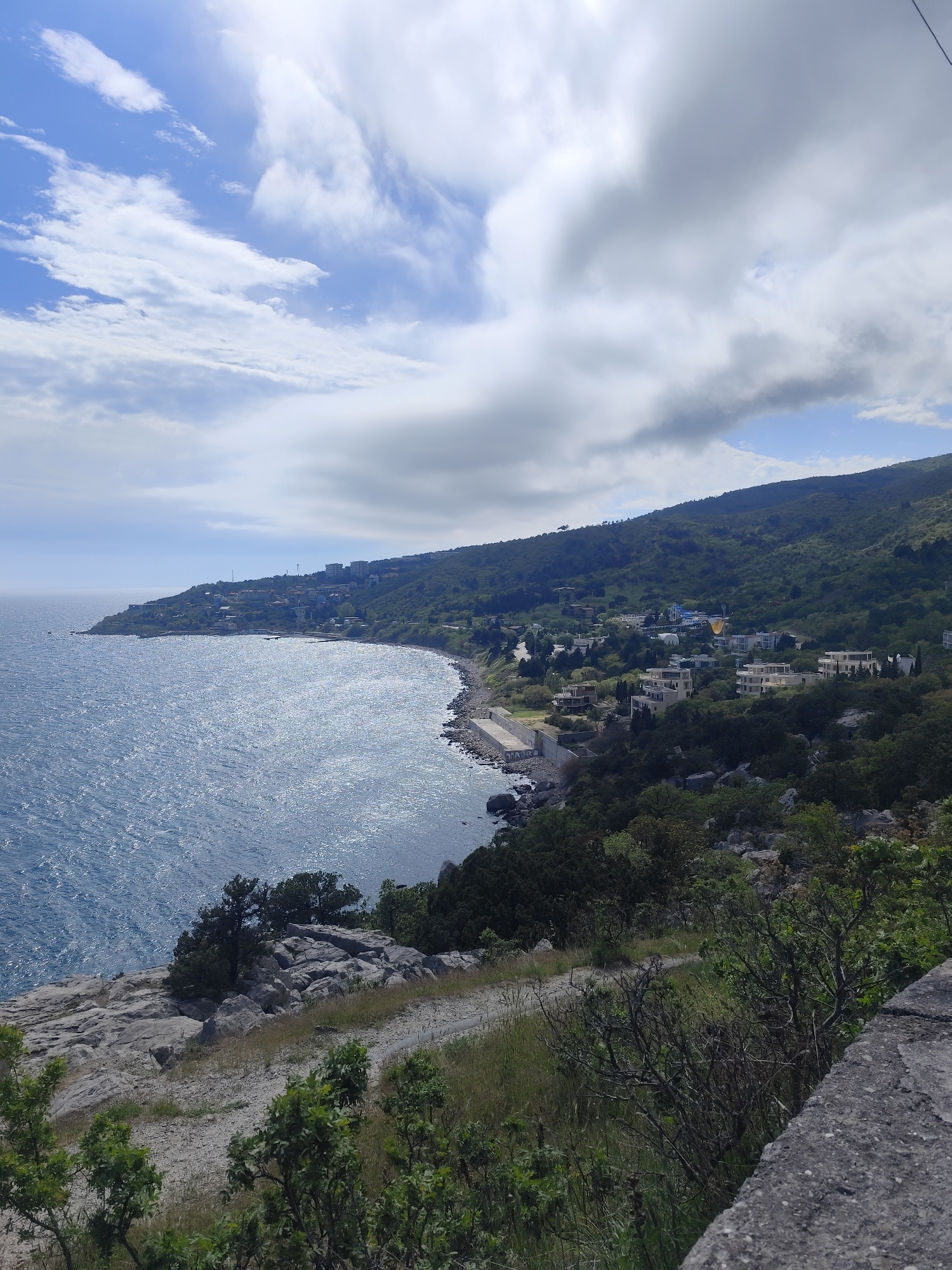 The image captures a coastal scene with a rocky shoreline meeting a blue sea under a sky with scattered clouds. A land formation covered in green vegetation and sparse buildings runs along the coastline. In the foreground, there are rocky surfaces and green bushes, suggesting a cliffside or elevated viewpoint. It is unknown what city or country this photo was taken in.