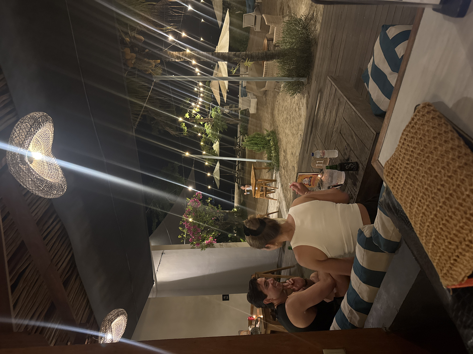 The image captures an outdoor dining scene at a restaurant or bar in Nusa Penida, Indonesia, during the evening. The environment features sandy ground with outdoor seating areas lit by string lights strung across palm trees. Visible are tables, chairs, and white sun umbrellas. Two people are seated at a table with drinks, and there are decorative straw lamps hanging from the ceiling. The scene suggests a relaxed, beach-side dining experience.