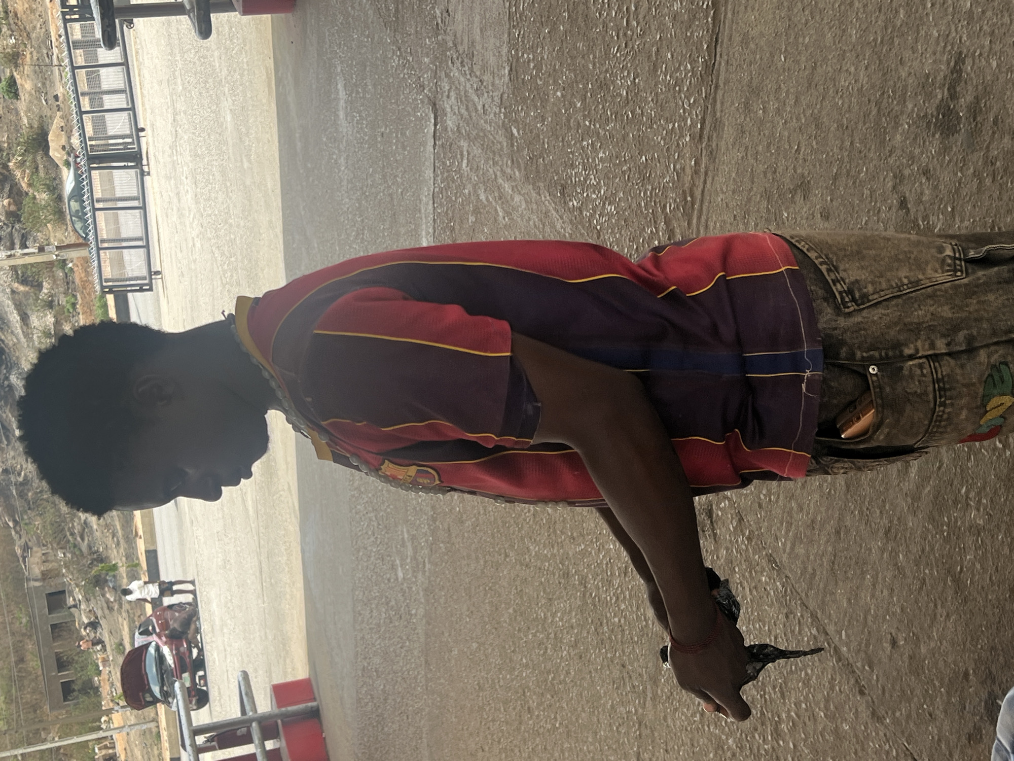 A person stands on a concrete surface in an outdoor setting, likely a public area within the city of Jos, Nigeria, judging by the visible landscape and structures in the background. The individual is wearing a patterned shirt and denim pants. They are holding a dark-colored object in their hands, with an unknown action taking place. The environment includes a view of distant buildings and possibly a hillside, suggesting an urban or semi-urban location within Jos. There is also evidence of a vehicle and street furniture in the scene.
