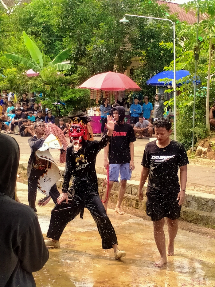 An outdoor cultural event is depicted on a wet, unpaved road in Gebyog, Indonesia, under bright daylight. In the foreground, a person partially obscured and facing away from the camera in a dark hooded garment observes the scene.
The central focus is a performance involving four individuals. A person in a black long-sleeved top and black pants, wearing a red demonic mask with white teeth and a dark, feathered headdress, stands in a wide stance with arms slightly extended, covered in water splashes. To their immediate left, another individual in dark attire holds a large, stylized white and brown prop resembling an animal head or a shield, also wet. To the masked figure's right, a young man in a black t-shirt with "BALIHAI" text and blue shorts stands barefoot, raising his right hand, heavily sprayed with water droplets. Further right, another young man in a black t-shirt with "JOKO REWANT LOKAL CULTURE" text and black shorts walks barefoot, also covered in water. Numerous water droplets are visible in the air across the midground.
In the mid-background, a crowd of spectators, primarily young men, are seated or standing on the left side of the road, observing the performance. On the right, a red market umbrella and a blue market umbrella are visible, suggesting vendor stalls. Lush green trees and foliage dominate the background, with a white street lamp extending over the scene from the right. The ground is visibly wet, indicating active water splashing during the event.