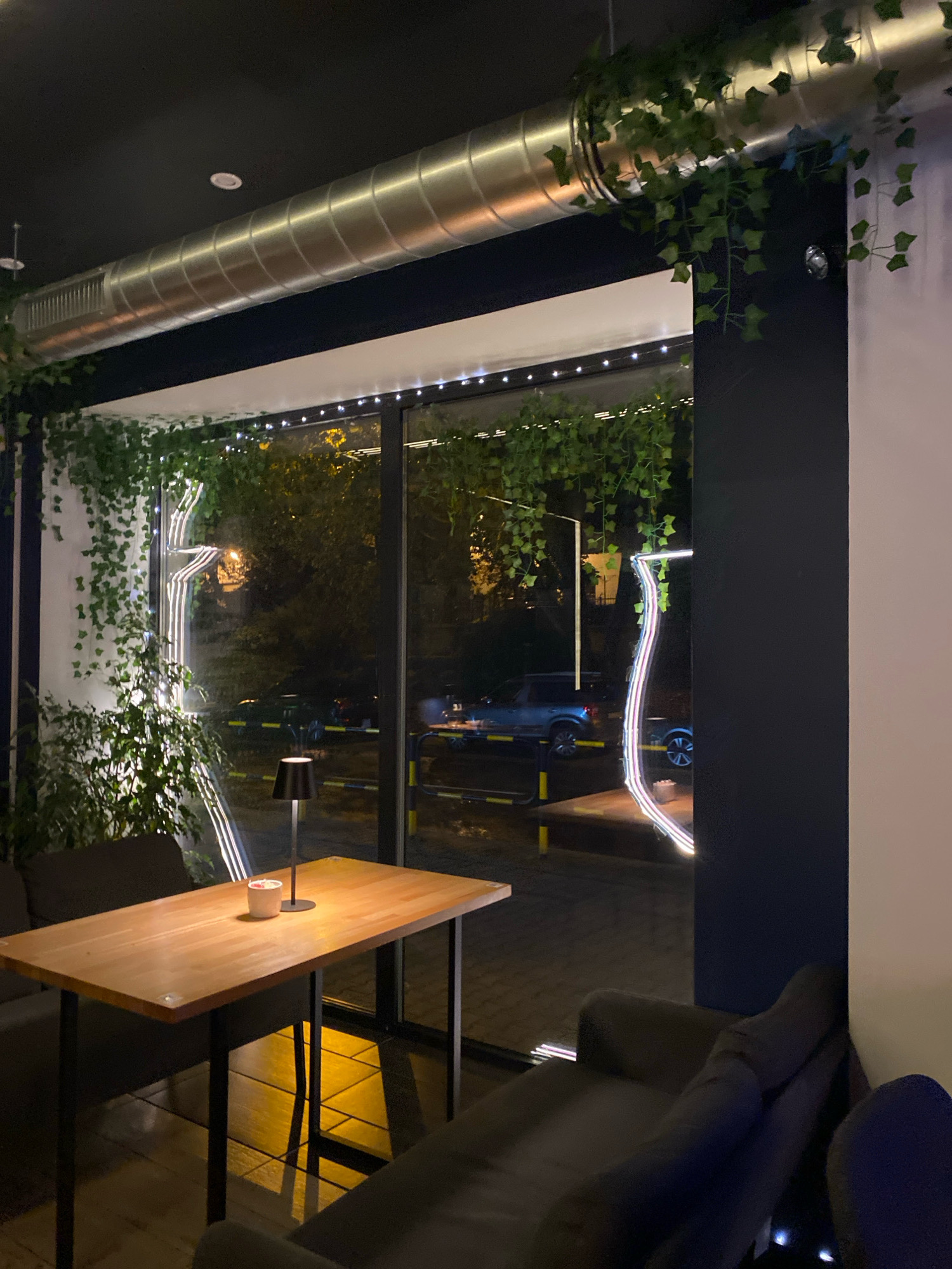 The image depicts the interior of a modern establishment, likely a cafe or restaurant, with a large window overlooking a street at night. Inside, a wooden table with black metal legs is positioned centrally, set with a small white pot. A black modern table lamp sits on the table, casting a warm glow. To the left of the table, a dark grey sofa is visible, and to the right, another sofa partially occupies the frame.
The window reveals a street scene with vehicles, including a blue car, parked or driving. The outdoor environment is illuminated by streetlights and the neon-like decorative lights inside the establishment, which are shaped like stylized glasses or vines. Artificial green ivy hangs from a large metal ventilation duct along the ceiling and from the frame of the window, creating a lush, indoor-outdoor aesthetic. The overall ambiance is dim and intimate, suggesting nighttime.
This scene is located in Gliwice, Poland. Specifically, the establishment is identified as "Kawiarnia Art Cafe," situated at ul. Rynek 15, 44-100 Gliwice. The cafe is known for its unique decor, which incorporates a significant amount of greenery and distinctive lighting, as evidenced by the image.