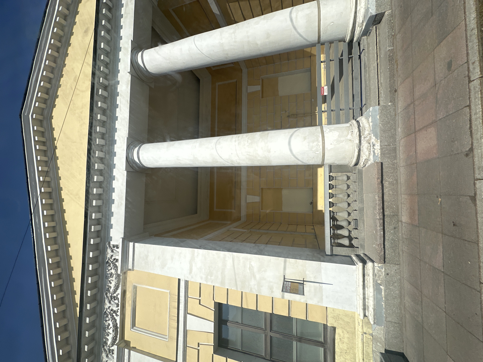 The image depicts a building in Saint Petersburg, Russia, featuring classical architectural elements. Two large cylindrical columns dominate the view, with a yellow facade visible behind them. The building's exterior is adorned with decorative details, including a window and intricate ornamentation. A sidewalk runs along the right side of the image, suggesting the building is located on a street. The sky is visible in the upper left corner, indicating a clear day.