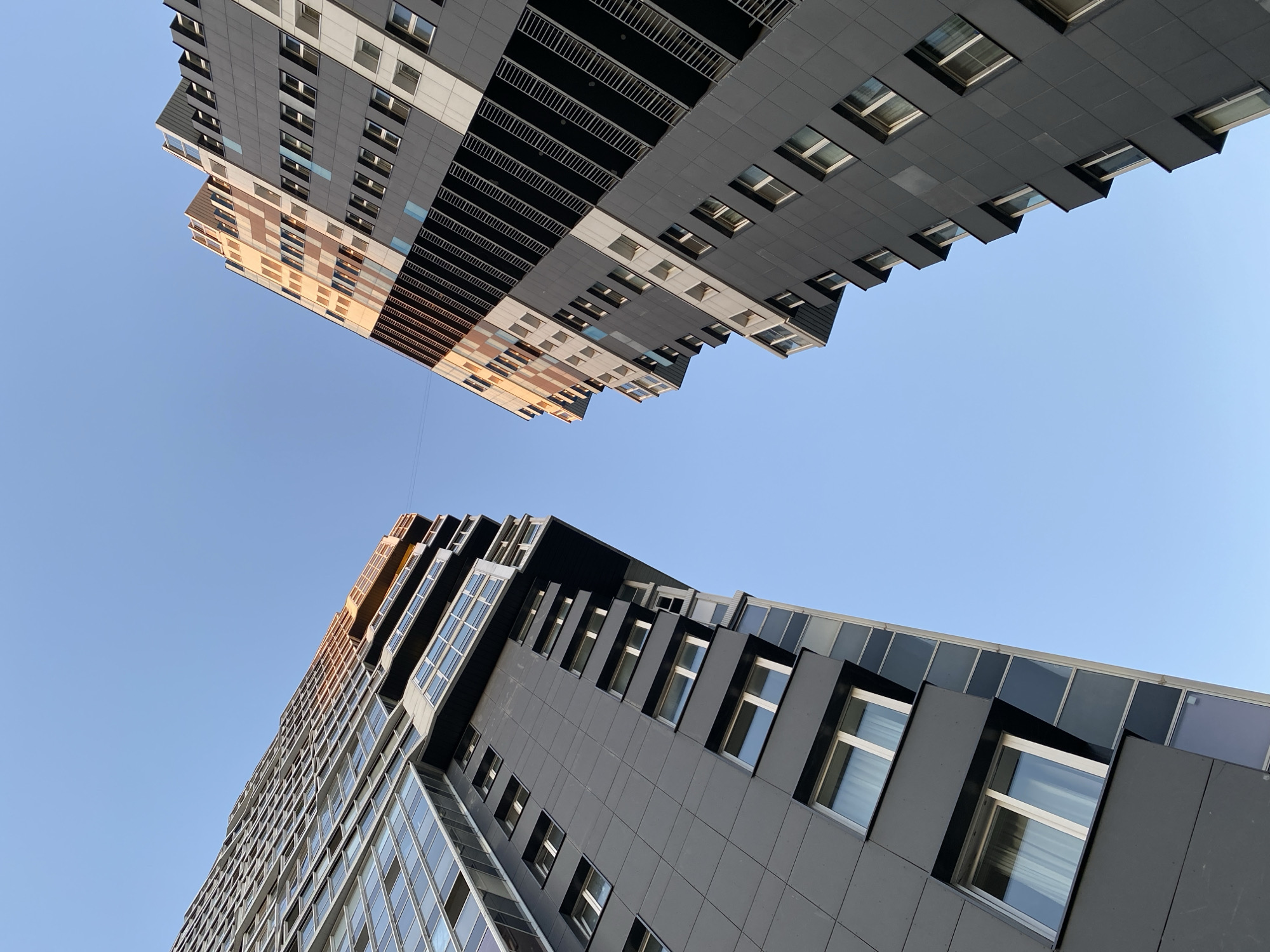 The image shows a low-angle shot of two modern high-rise buildings against a clear blue sky. The buildings are viewed from below, emphasizing their height and architectural design. One building on the left has a facade with alternating bands of light and dark grey panels and some sections with a checkered pattern of brown and beige. It has many windows, some of which are visible from this angle. The other building on the right is predominantly dark grey, with a more angular and stepped design. This building also features numerous windows, many of which appear recessed within dark frames, creating a layered effect. The sun is casting a warm glow on the upper portions of the buildings, suggesting it is either early morning or late afternoon. The sky is a uniform, cloudless blue, indicating clear weather. The architectural style of the buildings, with their sleek lines and modern materials, suggests an urban or developing suburban setting. The overall impression is one of contemporary urban architecture and the vastness of the sky. No people are visible in the frame. The location is identified as Kudrovo, Russia.