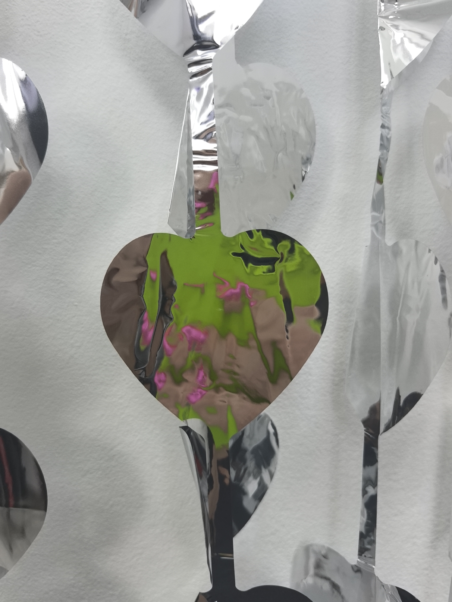 The image shows a close-up of silver, heart-shaped, metallic foil decorations hanging against a white wall. The decorations reflect the environment behind them, revealing glimpses of green foliage and pink flowers. The decorations appear to be part of a garland or string. The setting or specific location in the city of Chaikovsky, Russia is not evident from the image itself.