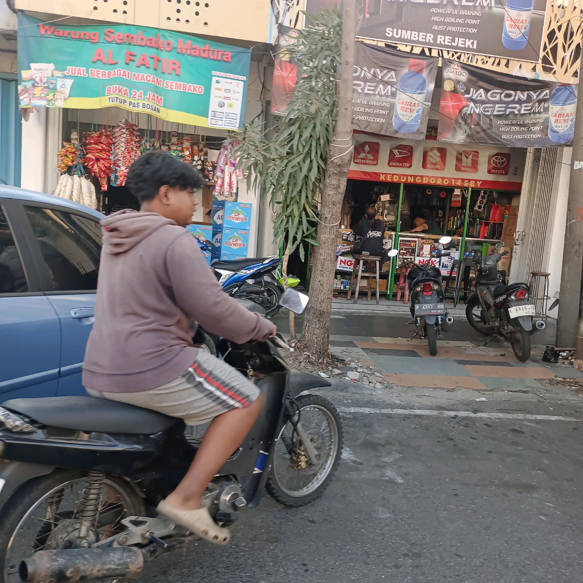 The image shows a street scene in front of a shop in Surabaya, Indonesia, specifically at KEDONGDORO 14 SBY. A young male, wearing a brown hoodie and plaid shorts, is riding a black motorcycle from left to right across the frame. He is wearing beige clogs. In the background, the shop displays several banners. One banner, in green and red, reads "Warung Sembako Madura AL FATIR JUAL BERBAGAI MACAM SEMBAKO BUKA 24 JAM TUTUP PAS BOSAN". Other banners advertise vehicle parts, featuring brands like Mitsubishi, Daihatsu, Suzuki, Isuzu, and Toyota. Two other motorcycles are parked in front of the shop. A blue car is partially visible on the left side of the image. The environment appears to be a typical street in an urban area, with some debris on the pavement.