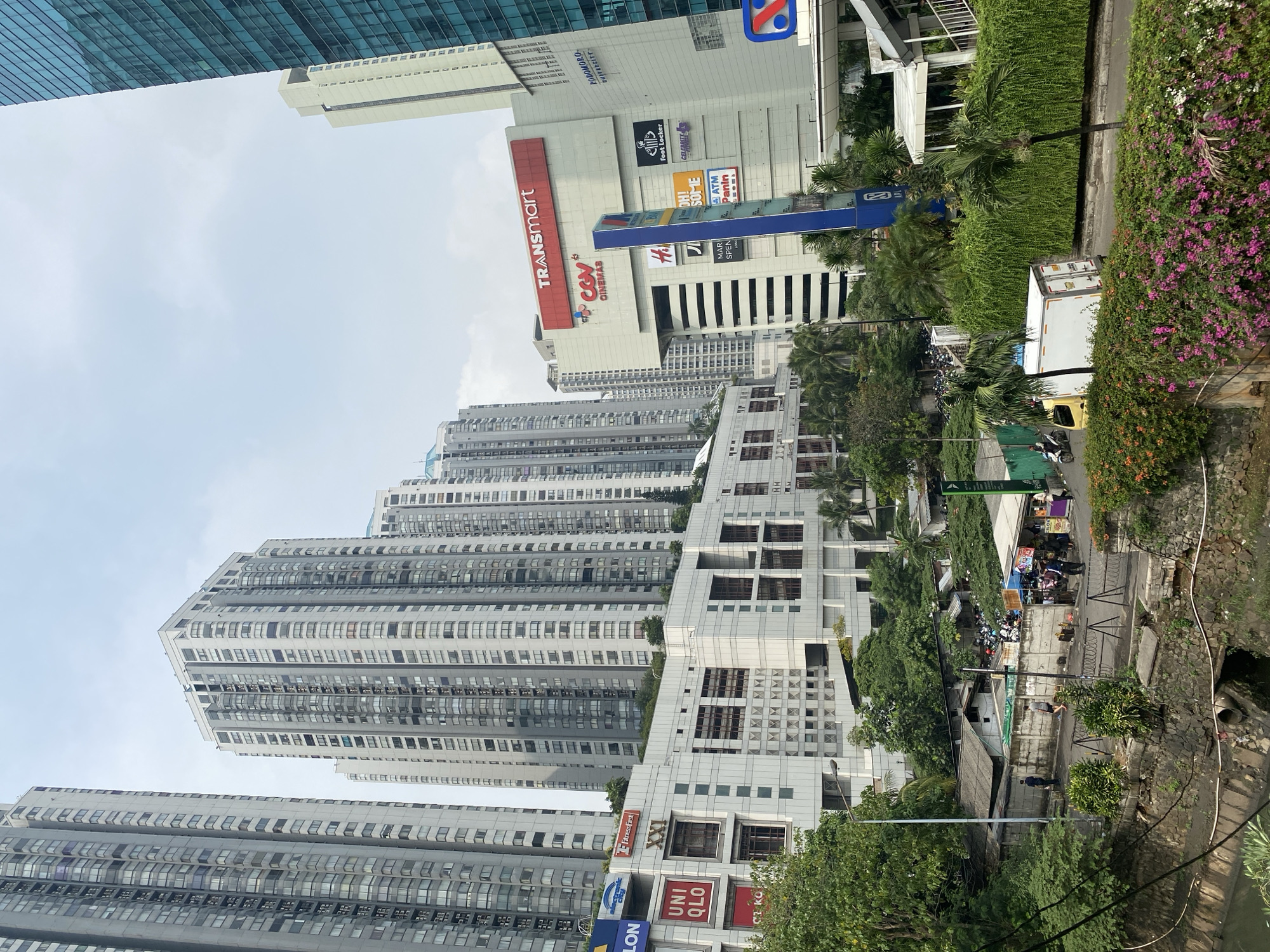 This image depicts a cityscape in West Jakarta, Indonesia, dominated by a cluster of tall, modern buildings. In the foreground, a canal runs along the bottom of the frame, bordered by a stone wall and lush green foliage interspersed with vibrant pink flowers. A dirt path runs alongside the canal, with several people walking and standing around. Beyond the path, a collection of smaller, low-rise buildings are visible, some with corrugated roofs.
Further back, a large, white, multi-story commercial building stands out, featuring prominent signage for "TRANSMART" and "CGV CINEMAS." This building is surrounded by other structures, including a tall, glass-fronted skyscraper on the right side of the frame, which is partially obscured. A bright blue advertising pole stands in front of the Transmart building. Various retail store logos, such as "UNI QLO," "Fitness First," and "Foot Locker," are visible on the facades of the buildings, indicating a commercial hub. The sky is overcast.
In the middle ground, a white truck is parked on a road. There are several motorcycles parked near the smaller buildings, and a few individuals are engaged in activity around them. The overall scene suggests a bustling urban environment with a mix of commercial and residential structures, typical of a developed area in a large city. The contrast between the modern skyscrapers and the more rudimentary structures in the foreground highlights different aspects of urban development.