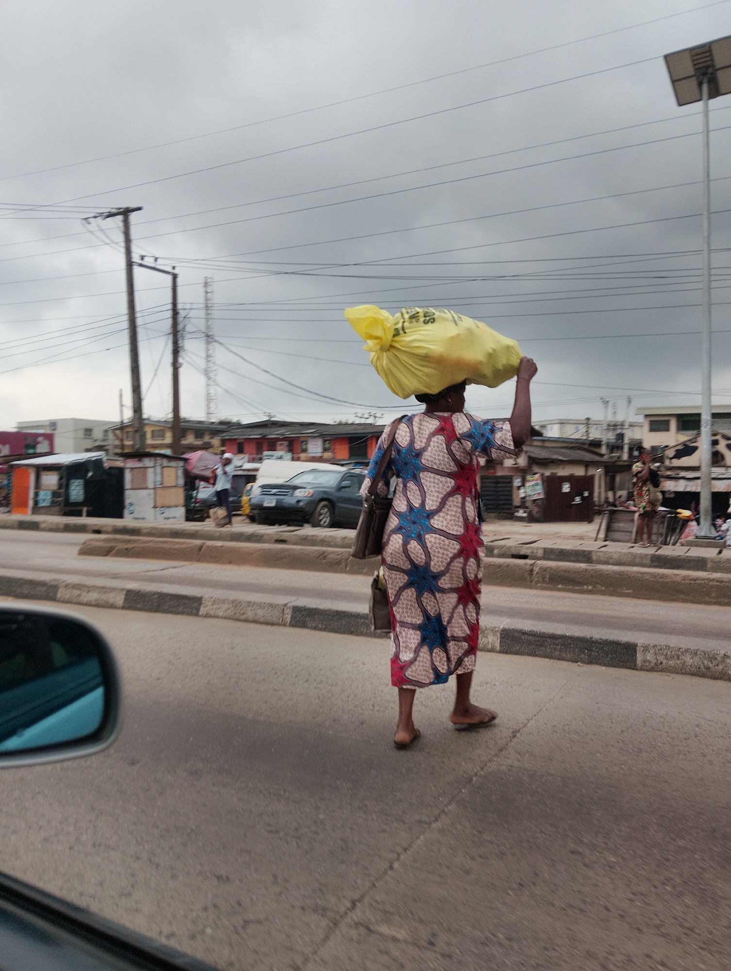 A woman walks along a paved road, carrying a large yellow bag of produce on her head. She is wearing a patterned dress and sandals. In the background, there are several market stalls and buildings, with power lines visible overhead. A dark-colored car is parked on the side of the road. The sky is overcast. This scene is consistent with the environment of Shangisha, Nigeria.