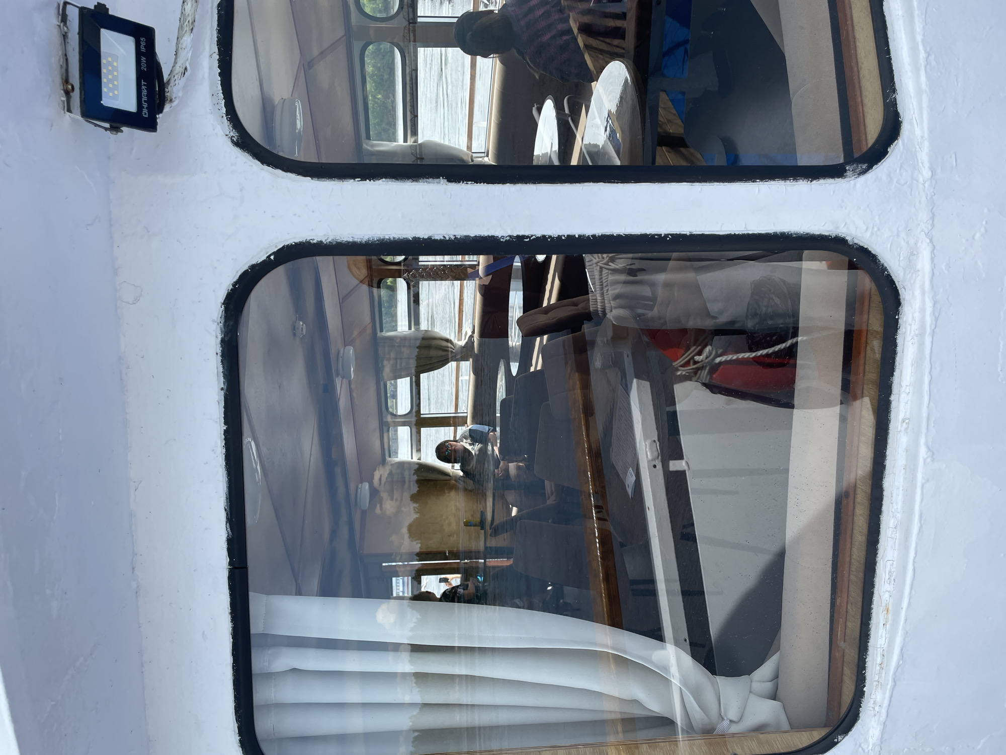 The image captures a close-up, exterior view of a white boat or vessel, oriented vertically, showcasing two stacked rectangular windows. Through the upper window, a person with dark hair, wearing a red and black plaid shirt, is visible from the waist up, appearing to be seated at a round table or operating equipment. Reflections of the surrounding water are also visible outside this window.
The lower window reveals another individual wearing glasses and a light-colored top, looking forward or to the side. Inside the cabin, visible through both windows, are wooden interior panels, brown upholstered seating, and white pleated curtains along the bottom edge of the lower window. The overall interior suggests a passenger area or salon.
The exterior of the boat is painted white, with some textured surfaces and signs of wear. Black sealant outlines both windows. To the upper left of the window frames, a black rectangular LED floodlight is mounted on the white hull. The visible text on the floodlight reads "ФОНАРЬ" (meaning "lantern" or "flashlight" in Russian), "20W," and "IP65."
The bright light and reflections on the water indicate that the scene takes place during the daytime. Given the location context of Perm, Russia, the vessel is likely navigating the Kama River or a similar body of water.