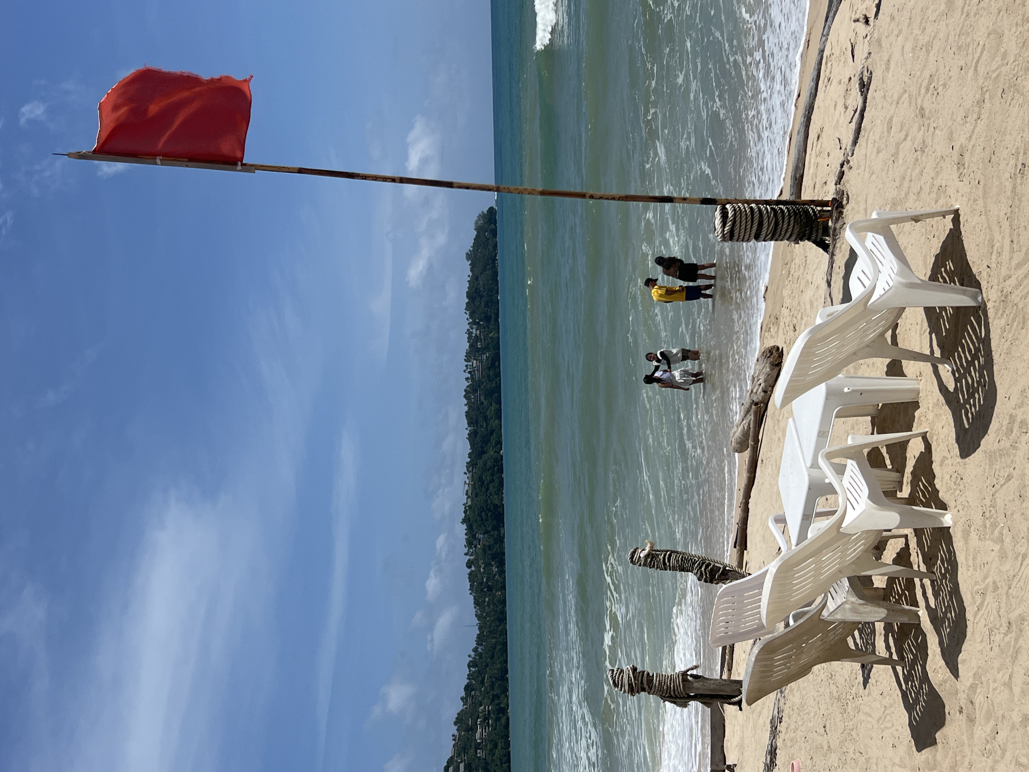 A red flag flies on a pole on a sandy beach in Choeng Thale, Thailand. The flag indicates caution or danger, likely due to strong currents or rough seas. Several white plastic chairs and a table are set up on the sand in the foreground, casting shadows. In the background, a group of four people are wading in the shallow water near the shore. The sea is a turquoise green with visible waves, and a lush, green hillside with buildings is visible in the distance across the water. The sky is blue with scattered white clouds. Two wooden posts wrapped with rope stand in the shallow water to the left of the image.