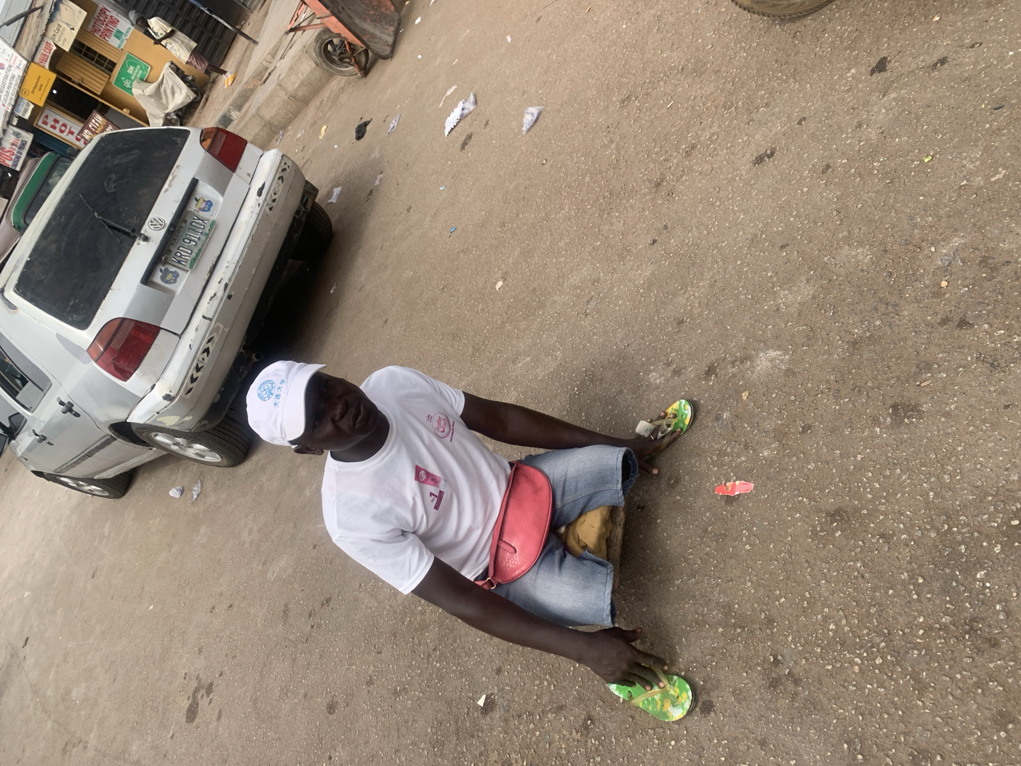 A man is sitting on the ground next to a gray Volkswagen Golf with the license plate KRD 911DX. The man is wearing a white t-shirt, denim shorts, a pink waist bag, a white hat with Chinese characters, and green and yellow sandals. The t-shirt features the phrases "大连大学," the number "1," and "Feather Touch." The environment consists of an asphalt street with scattered debris and a building with signs indicating services such as "POSTAL," "CHARGING OF PHONES," "REGISTRATION," "COLOUR PRINTING," "ONLINE REDISTRATION," "PHOT," "CYBER CAFE SIM Card," "PHOTOCOPY PRINTING," and "glo SIM." The location is Lagos, Nigeria.