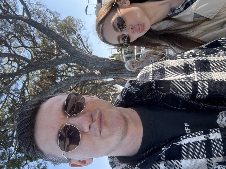 A selfie-style photograph depicts three individuals outdoors in Peschiera del Garda, Italy, under clear daylight conditions. In the immediate foreground, a male subject with short, dark hair, wearing gold-rimmed polygonal sunglasses, a black t-shirt with "OFF" partially visible, and a black and white plaid shirt-jacket, looks directly at the camera with slightly pursed lips. To his right and slightly behind him, a female subject with long dark hair, wearing narrow oval gold-rimmed sunglasses and a light-colored trench coat over a plaid shirt, looks towards the camera with her lips pursed in a pronounced kiss face. In the mid-ground, centered behind the foreground subjects, a second female with blonde hair, dressed in a light brown jacket, stands holding a white smartphone vertically in her right hand, gazing down at the device. The background features several large trees with substantial trunks and sparse foliage, characteristic of pines. Distant light-colored buildings with rooftops and a streetlamp are visible on the right, beneath a clear blue sky.