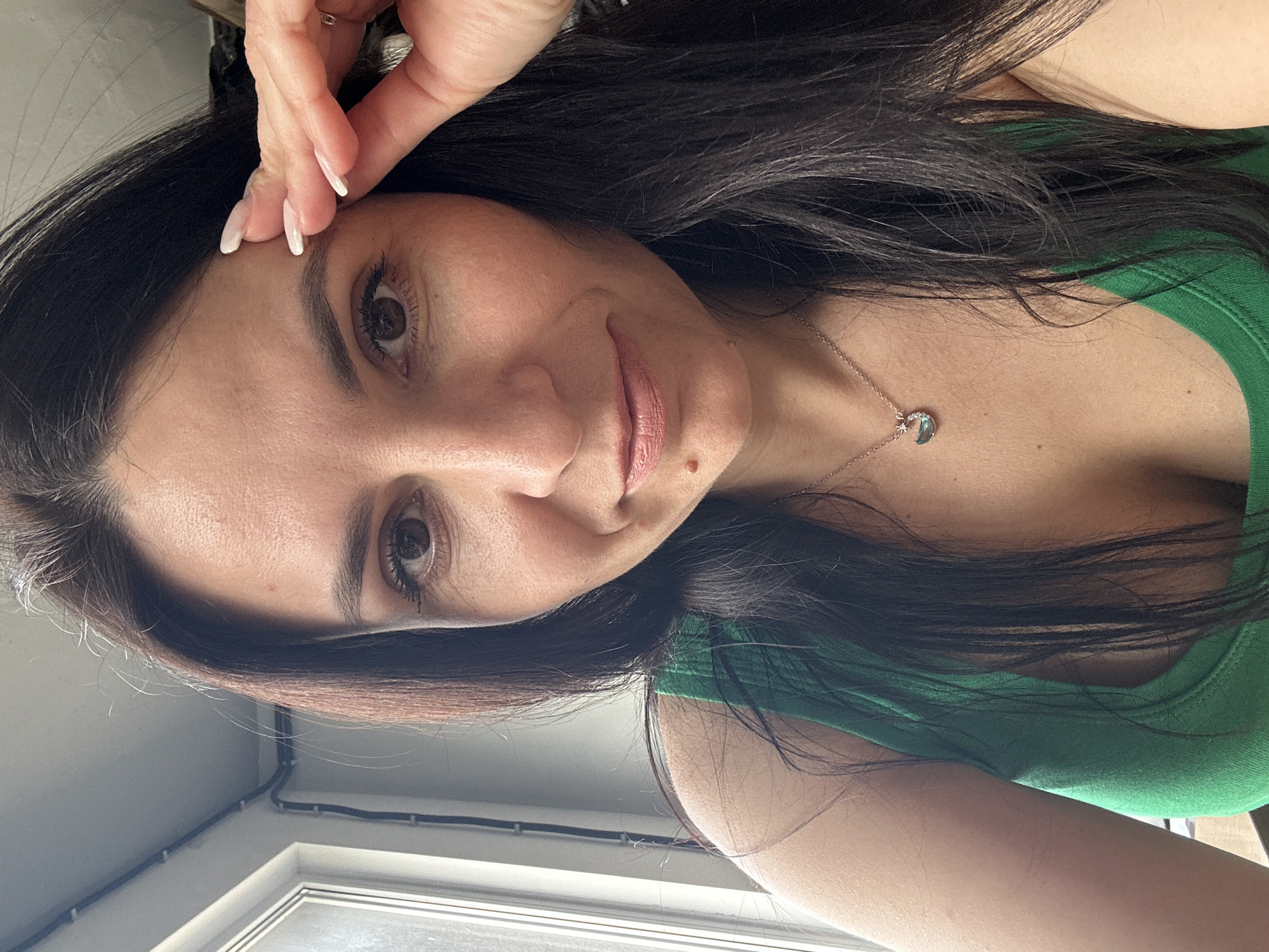 A woman with long, dark hair is looking at the camera. She is wearing a green top and a necklace with a circular pendant. Her left hand is raised to her forehead. The background shows a window and a white wall with some electrical wiring. The location is Unknown, Unknown.
