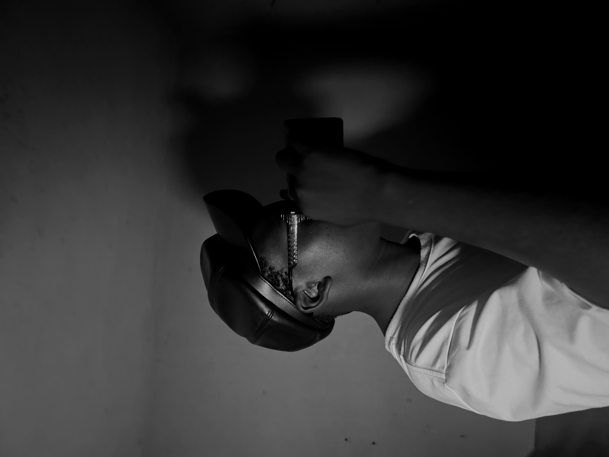 A monochrome medium shot depicts a person pouring liquid over their head from a phone. The individual is wearing a cap and a light-colored shirt. The location appears to be indoors, with a wall in the background. No clear indication of the city of Asaba, Nigeria, is discernible from the image.
