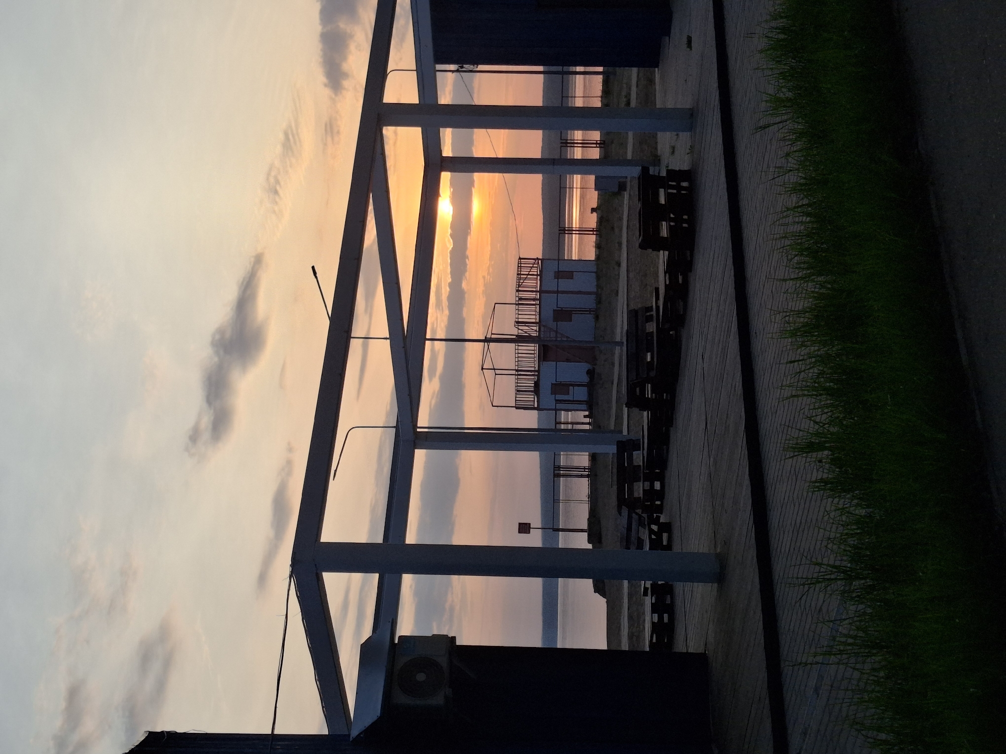 The image depicts a sunset scene in Chaikovsky, Russia. A metal structure dominates the foreground. Through the structure's frame, a beach with various buildings is visible. The sky is a mix of orange and grey, indicating either sunrise or sunset. Below the structure, there are dark objects, possibly tables or chairs. To the right of the frame, there is a green strip of grass and a paved walkway. The image is slightly rotated.