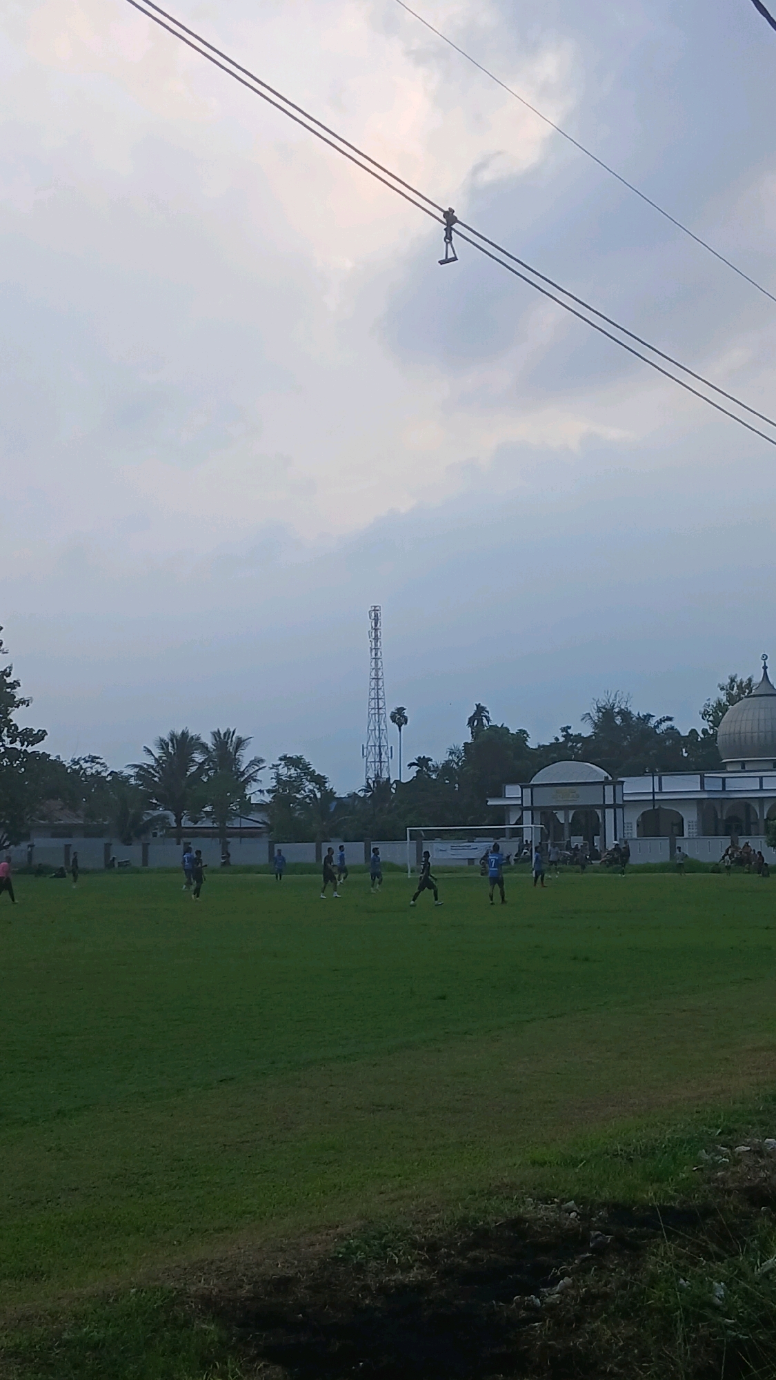 In the city of Sunggal, Indonesia, an outdoor football game is underway on a lush green field. The sky is overcast, with a prominent telecommunications tower visible in the distance. To the right of the tower, a white building with a dome, likely a mosque, is situated. Several individuals, appearing to be players, are scattered across the field, some engaged in the game, while others are standing. Overhead, two thick cables are strung across the sky, one of which has a small apparatus attached. The foreground of the image features a dark, overgrown area of vegetation.