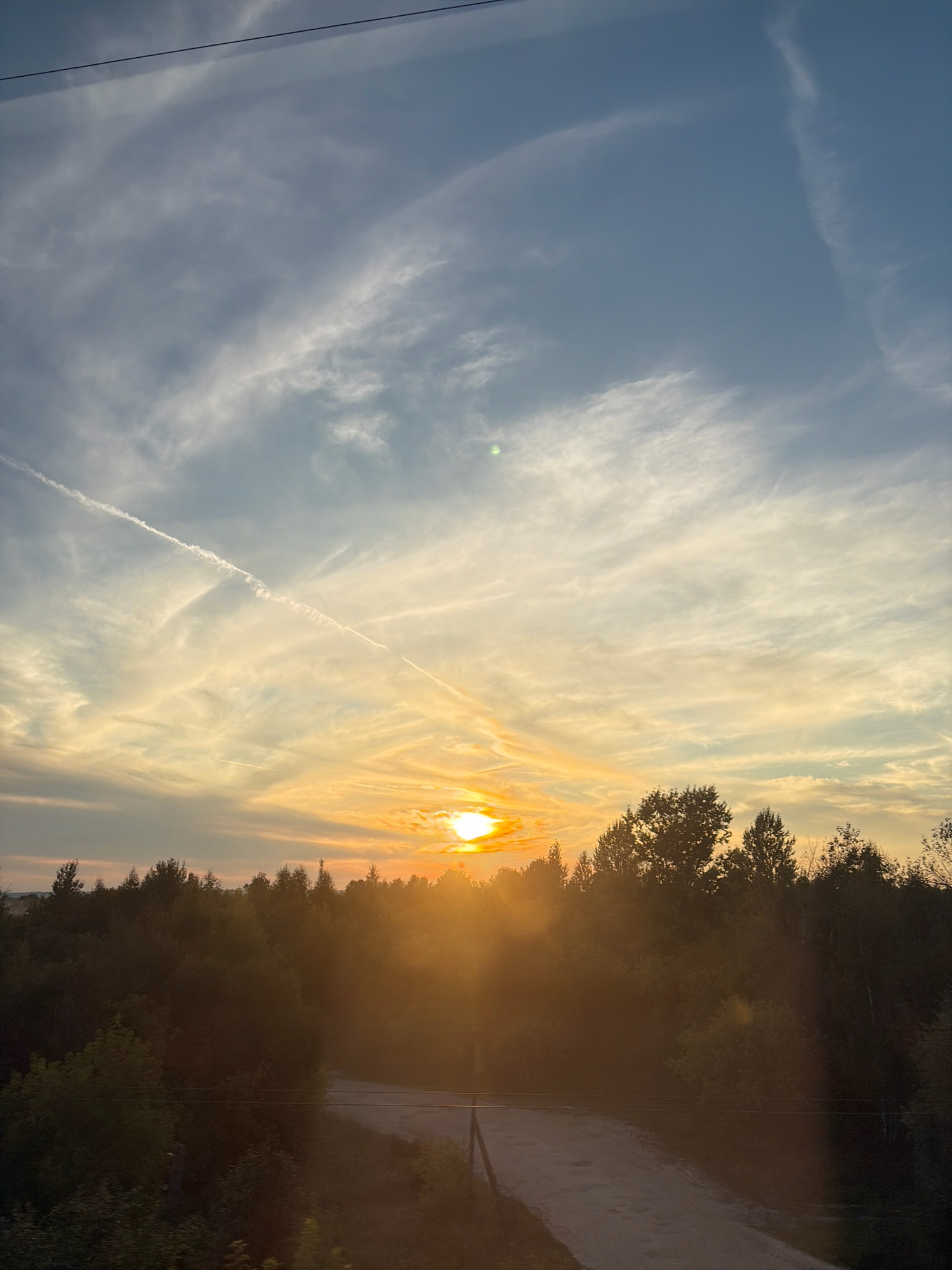The image depicts a sunset over a rural landscape, likely in the vicinity of Галицы, Russia. The sun is low on the horizon, casting a warm, golden light across the sky and a dense line of trees that forms the foreground. Wispy clouds are scattered across the sky, some illuminated by the setting sun and others tinged with blue. Contrails from airplanes are visible in the upper portion of the sky, indicating air traffic. A dirt road or path winds through the trees, disappearing into the shadows. Power lines stretch across the scene, adding a touch of human infrastructure to the natural environment. The overall impression is one of tranquility and the end of a day in a natural setting.