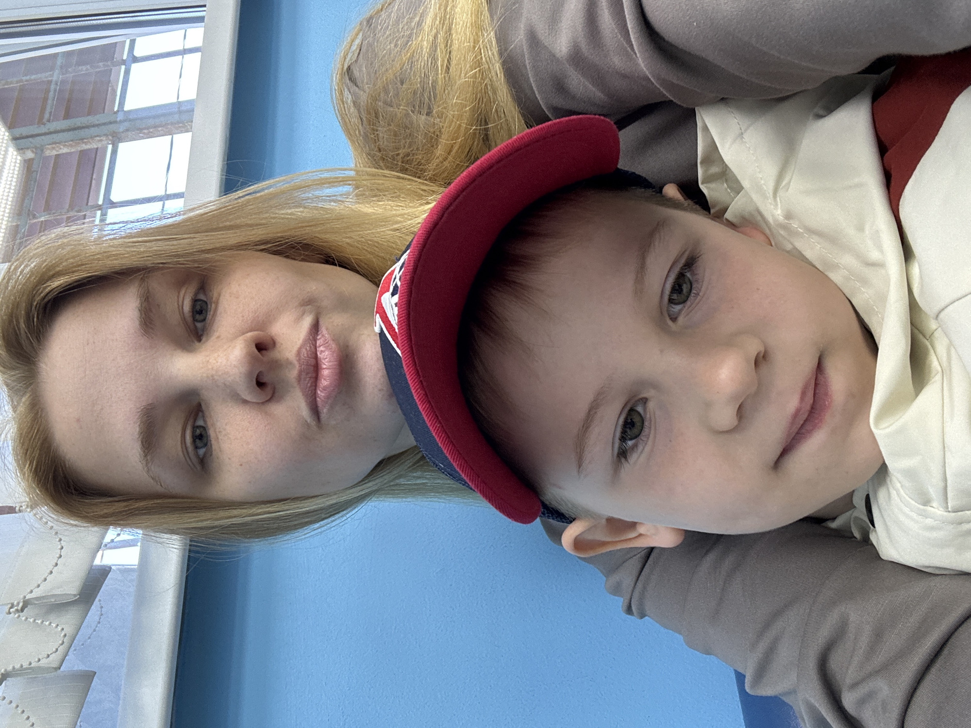 An adult female and a young male child are depicted in a close-up, head-and-shoulders portrait within an indoor environment in Rusanovo, Russia. The female, positioned behind the child, has long blonde hair, light-colored eyes, and visible freckles on her nose and cheeks. She wears a plain grey long-sleeved garment and looks directly at the camera with a slight pouting expression. Her left arm is visible, seemingly around the child. The child, a boy, appears to be approximately five to seven years old. He wears a blue baseball cap with a red brim and a partial white logo, a cream-colored hooded jacket over a red garment, and has light brown hair and greenish-brown eyes. He looks slightly to the viewer's left, exhibiting a subtle, closed-mouth smile. The background features a solid light blue wall in the lower and central sections. Above the blue wall, a window with white horizontal blinds is visible, allowing a bright, indistinct exterior view through its panes. The lighting suggests natural illumination originating from the window.
