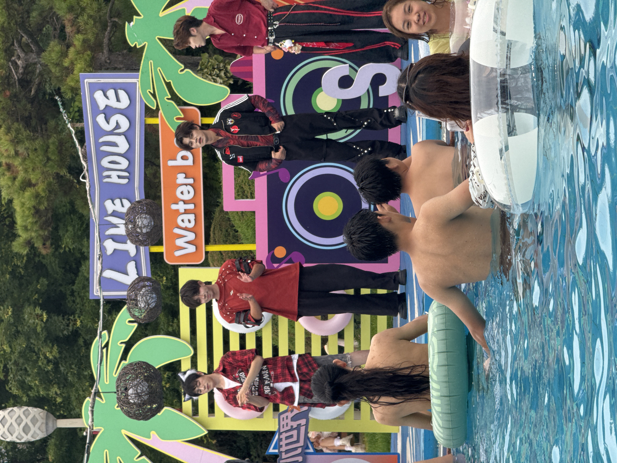 This media file shows a group of young people posing for a photo in an outdoor setting, possibly a pool party or event. The subjects are predominantly young Asian males, some of whom appear to be in a K-pop style group given their fashionable attire and coordinated poses. They are dressed in a mix of casual and stylish outfits, including jackets with intricate designs, plaid shirts, and dark pants. One individual wears a red jacket with black accents and studs, another a red and black plaid shirt, and a third a red shirt with similar embellishments.
The setting appears to be a vibrant outdoor area with colorful decorations. Behind the individuals, there are large, stylized cutouts of palm trees, and a brightly colored backdrop featuring abstract shapes and circles. Signs are also visible, one with blue text reading "LIVE HOUSE" and another with orange text that appears to say "Water b". The presence of inflatable rings, one white and one light green, and the shimmering blue water in the foreground strongly suggest a pool or a water-themed area. The water is clear and reflects light, indicating it is likely daytime. The overall mood seems to be festive and energetic, with the subjects looking engaged and some smiling. The scene is well-lit, suggesting a sunny day. The location context of Taihe District, China, provides a geographical anchor for the scene.