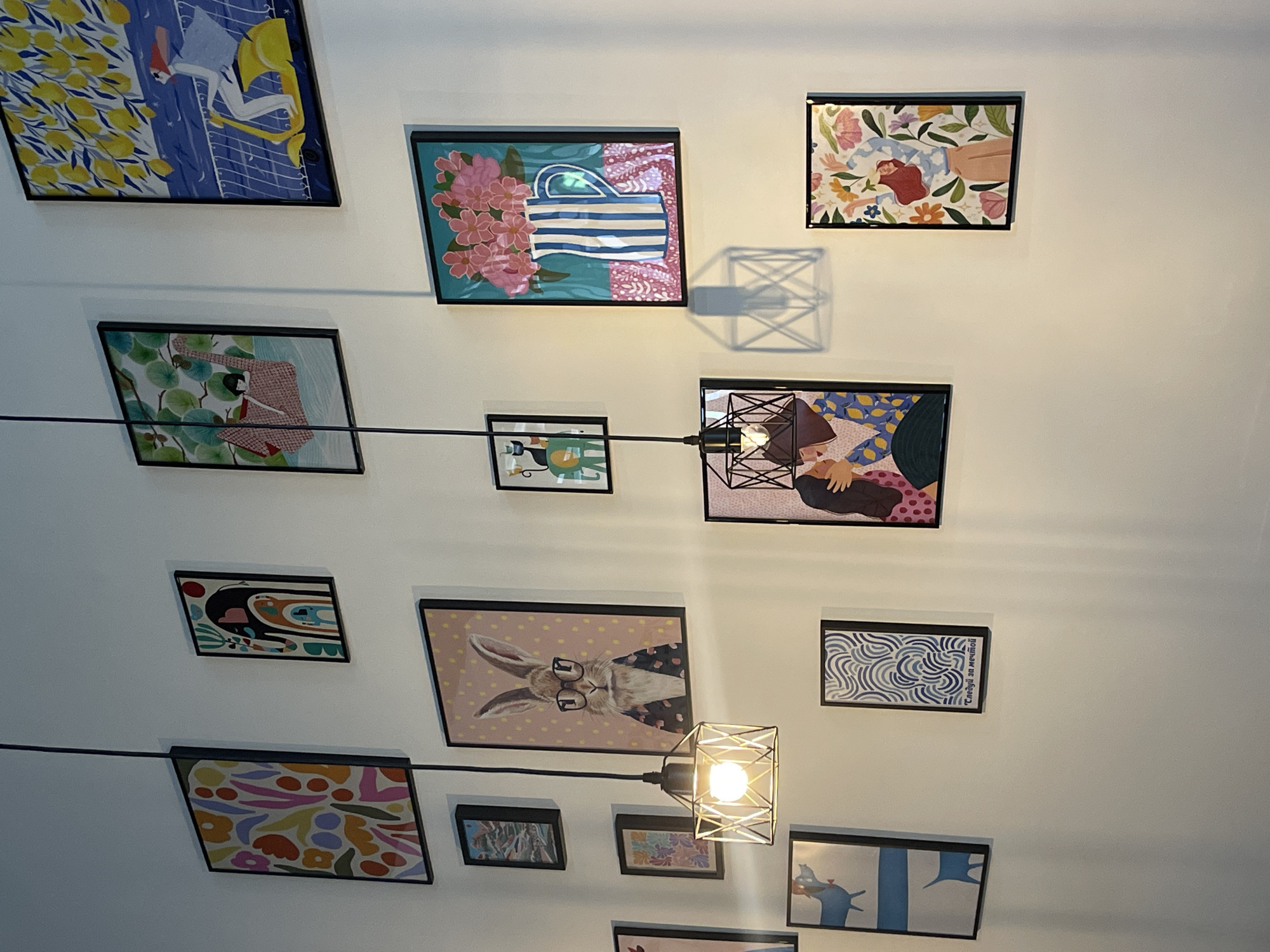 The image displays a collection of framed artworks hung on a white wall, likely in a domestic or commercial interior. The artworks feature a variety of styles and subjects, including abstract patterns, a depiction of a rabbit wearing glasses, a still life of flowers in a vase, a scene with a woman riding a scooter, a depiction of a couple embracing, and a framed print with blue wavy lines and Russian text that reads "Следуй за мечтой" (Follow your dream). Two pendant lights with geometric metal shades hang from the ceiling, illuminating the wall and casting shadows. The overall impression is one of a curated display of decorative art. The specific location of Donskoye, Russia is not discernible from the visual information provided in the image.