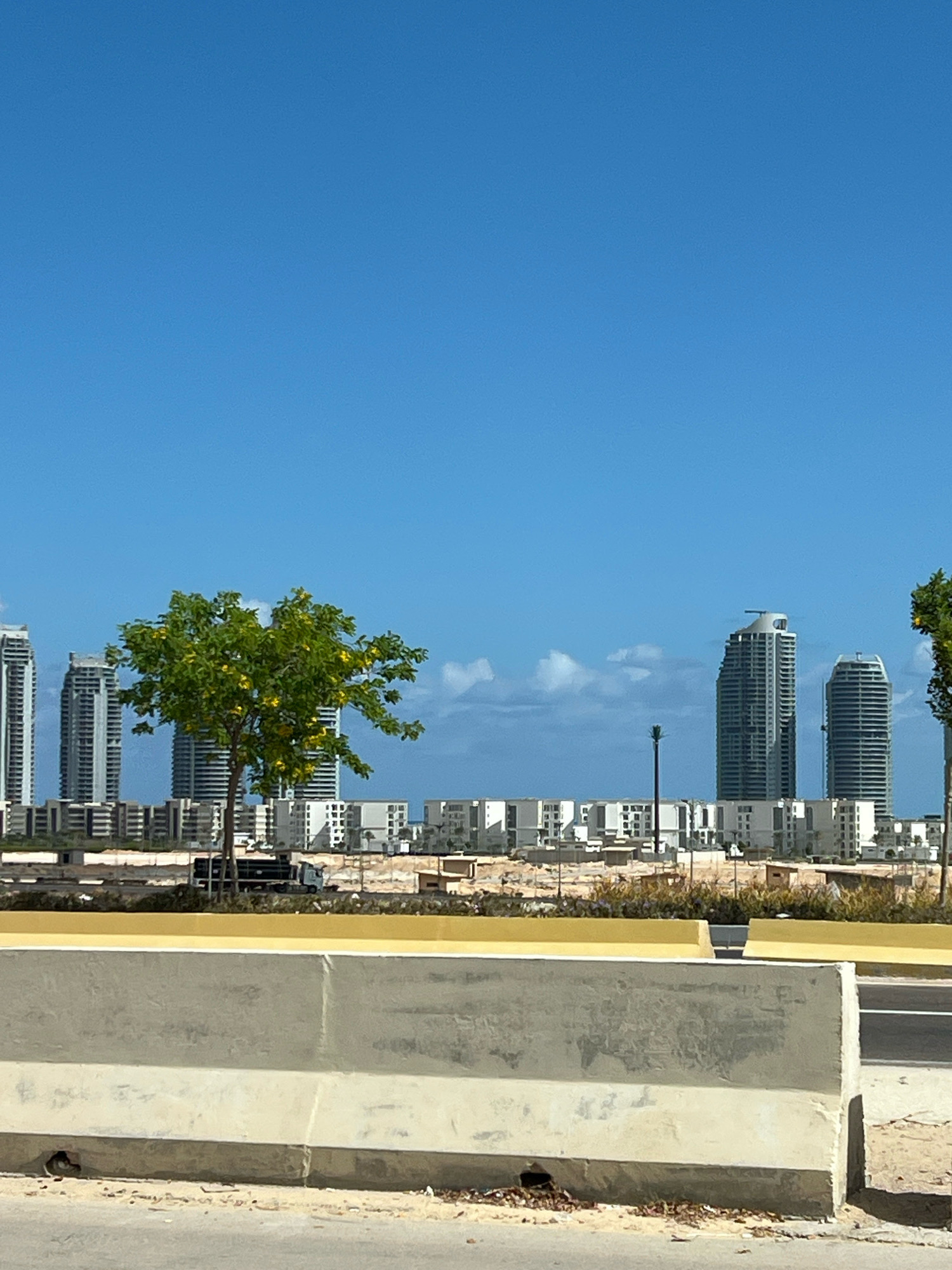 In the city of Sidi Abdel Rahman, Egypt, a wide shot captures a cityscape under a clear blue sky with sparse clouds. In the foreground, a concrete barrier with yellow accents runs horizontally, separating the viewer from the scene beyond. Behind the barrier, a dirt road leads towards a construction zone. A single green tree with yellow flowers stands prominently in the mid-ground, its branches reaching towards the sky. Beyond the tree, a cluster of modern, multi-story buildings form a distinct skyline. Further in the distance, several tall skyscrapers dominate the horizon, their sleek designs suggesting a contemporary urban environment. The overall scene depicts a developing area with a blend of natural elements and burgeoning infrastructure.