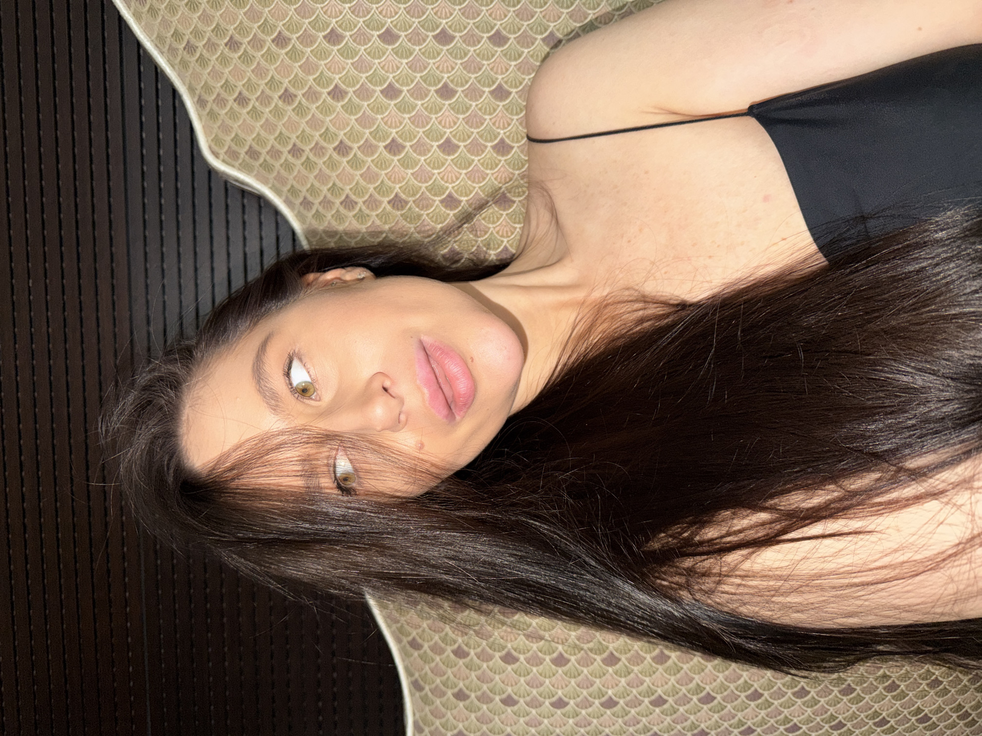 A young woman with long, dark brown hair is captured in a close-up portrait, lying down indoors. Her head rests on a patterned pillow, displaying a muted scale-like or scallop-shell design in shades of green, beige, and purple-brown. To the left of her head, a dark, vertically-panelled surface, possibly a headboard or wall, is visible.
The woman's face is turned towards the viewer, with her light-colored eyes (appearing hazel or greenish-brown) looking directly at the camera. Her full, pink lips are slightly parted, suggesting a pensive or contemplative expression. She is wearing a simple black top with thin straps. Direct sunlight illuminates her face and hair, creating strong highlights and revealing freckles on her neck and chest, indicating a daytime setting. The scene conveys a calm and intimate atmosphere, likely within a home in Khimki, Russia. No text is visible in the image.