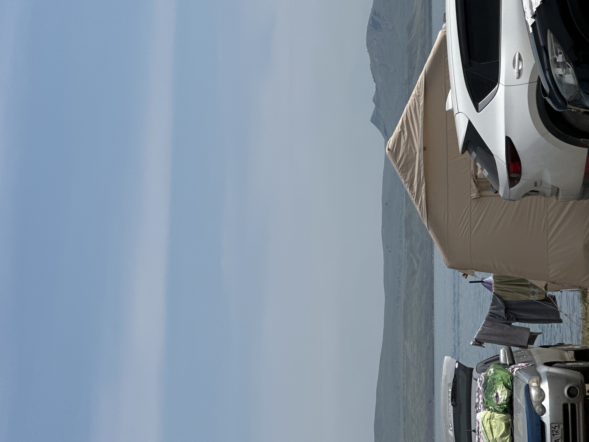 The image depicts a campsite near a body of water, possibly a lake or river, with hills in the background. Two cars are parked, one silver and one white. The silver car has its trunk open and several bags are piled on top of it. Clothes are hanging on a line strung between the silver car and a large tan tent. The white car is parked next to the tan tent, and the front of a black car is visible in the lower right corner of the image. The sky is clear with some faint clouds. The scene suggests a recreational or camping activity. The specific location of Колодезный, Russia, is not explicitly visible or indicated in the image.