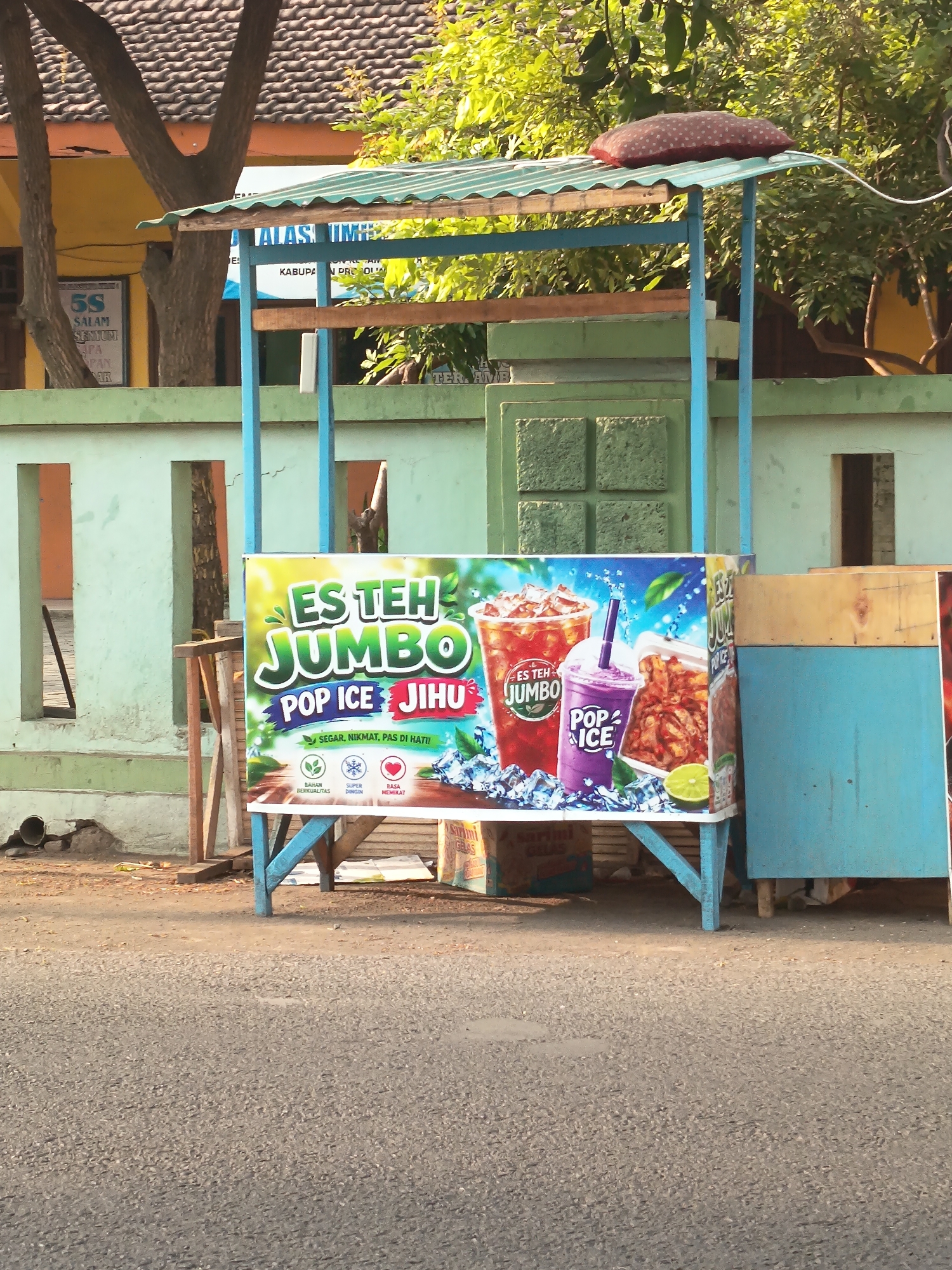 A street-level, eye-level outdoor shot shows a mobile beverage stall on an asphalt road. The stall features a green corrugated metal roof supported by blue wooden posts, with a wooden counter displaying a large advertisement. The advertisement prominently features the text "ES TEH JUMBO" in green and white, followed by "POP ICE JIHUI" in blue and red. Images of two drinks, one red with ice and a straw, labeled "ES TEH JUMBO," and a purple drink labeled "POP ICE," are showcased, along with a depiction of french fries and a slice of lime. Below the main text, smaller text reads "SEGAR, NIKMAT, PAS DI HATI!" and icons representing "BAHAN BERKUALITAS," "SUPER DINGIN," and "RASA MEMIKAT." The stall is situated in front of a light green wall with two rectangular openings and a darker green tiled section above. A partially visible blue wooden cabinet is attached to the right side of the stall. In the background, trees with green foliage are visible, and a yellow building with a dark tiled roof can be seen. The scene appears to be located in Kandangjati Wetan, Indonesia, as indicated by signage in the background reading "KABUPATEN PRO."