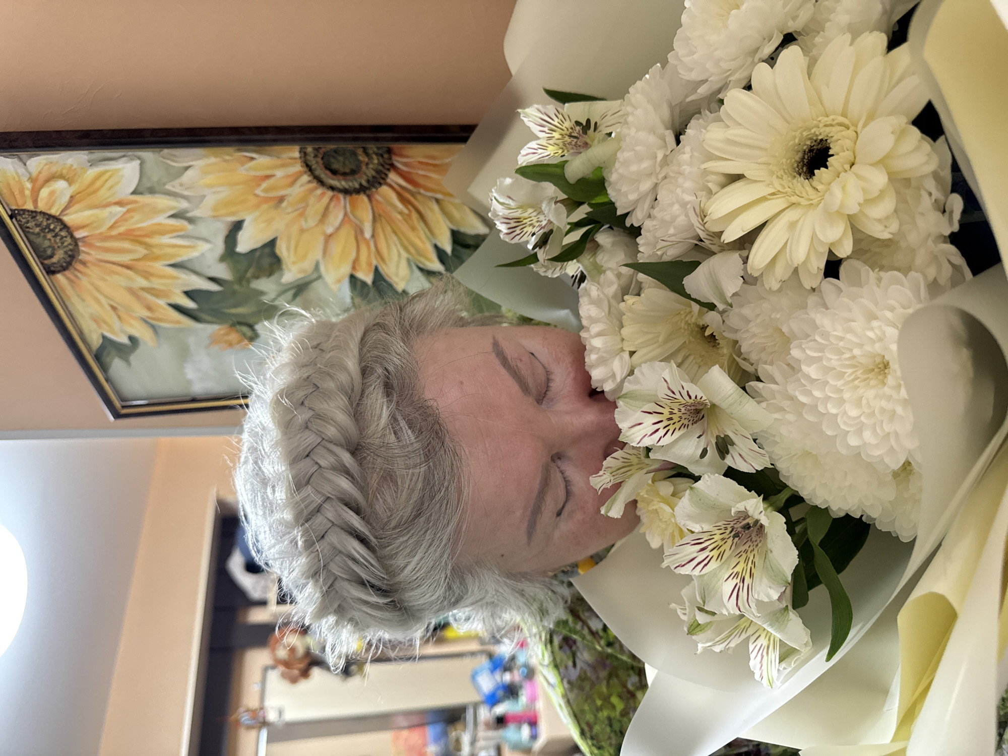 A woman with gray braided hair is holding a bouquet of white flowers to her face. A painting of sunflowers is visible on the wall behind her. The location is likely Chaikovsky, Russia.
