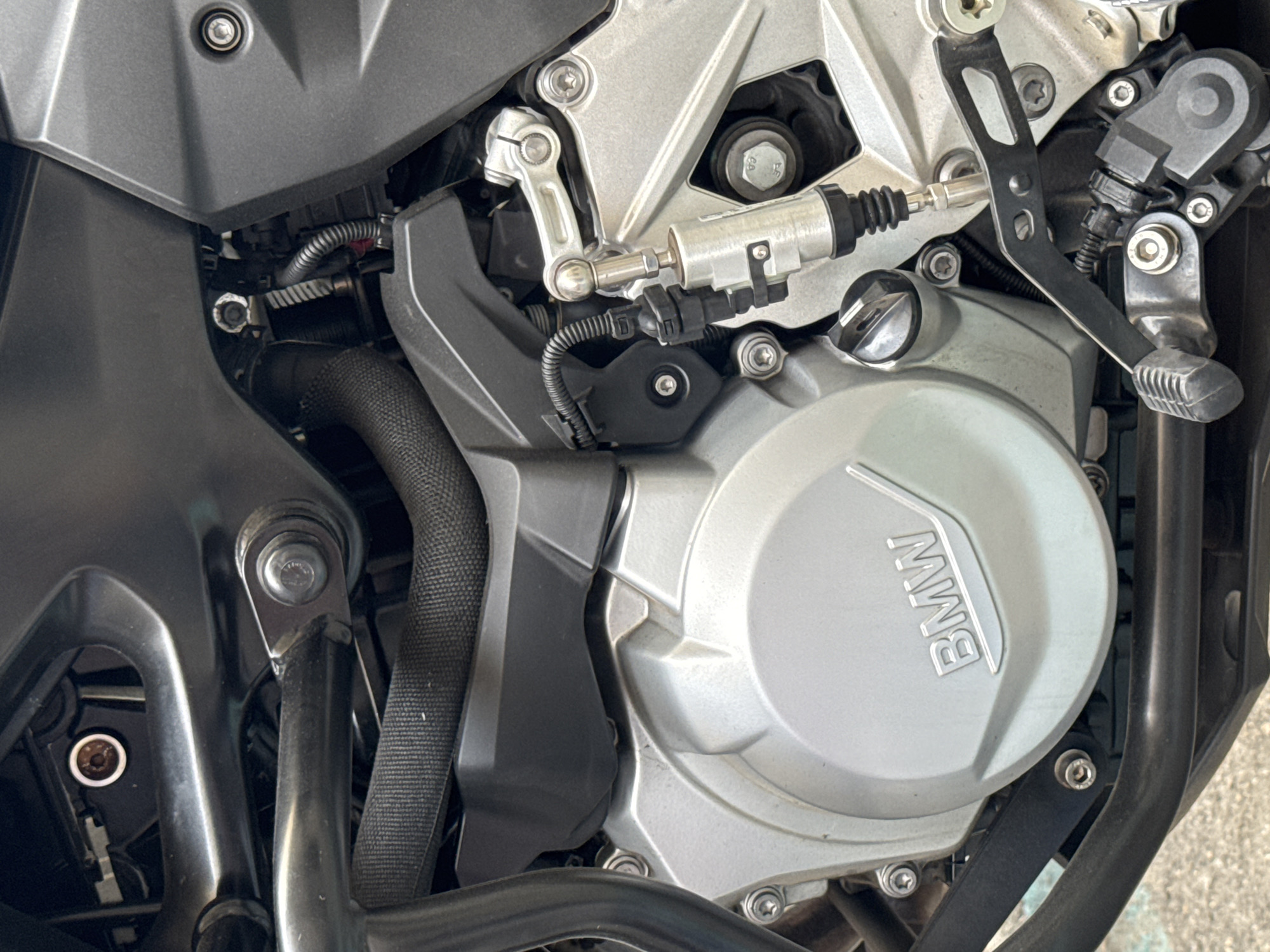 This close-up, high-angle shot shows a section of a motorcycle's engine. The engine casing is predominantly silver, with the "BMW" logo clearly visible on a circular cover. Various mechanical components, including levers, cables, and fasteners, are visible. A black protective sleeve covers a portion of a hose or cable. The overall setting appears to be an outdoor environment, possibly a street or parking area in Madrid, Spain, suggested by the lighting and potentially the surface texture in the background. There is no activity or event taking place; the image is a static capture of the motorcycle part. The lighting is bright, suggesting it is daytime. No people are visible in the frame.