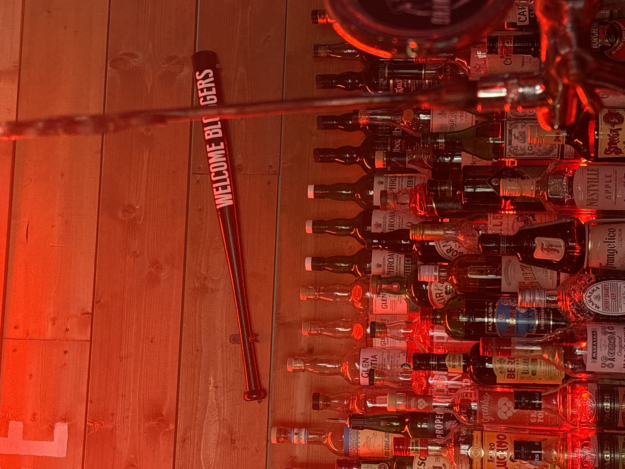 The image shows a bar with a wall display of liquor bottles. A baseball bat with the words "WELCOME BLOGERS" is on the left side of the image. The bar is located in Saint Petersburg, Russia. The bottle labels visible include "400 CONEJOS", "ESCO", "AMIKRO LUCANO", "PROPER", "Greenbar", "GLE BALT", "GLEN TIA", "GLEN", "BERO", "BERGIA", "COURVO", "NESTVILLE APPLE", "rangelico", "Ibeg be", "PHROA", "LE", "MARASKA", "ARD", "CLA", and "CAR".