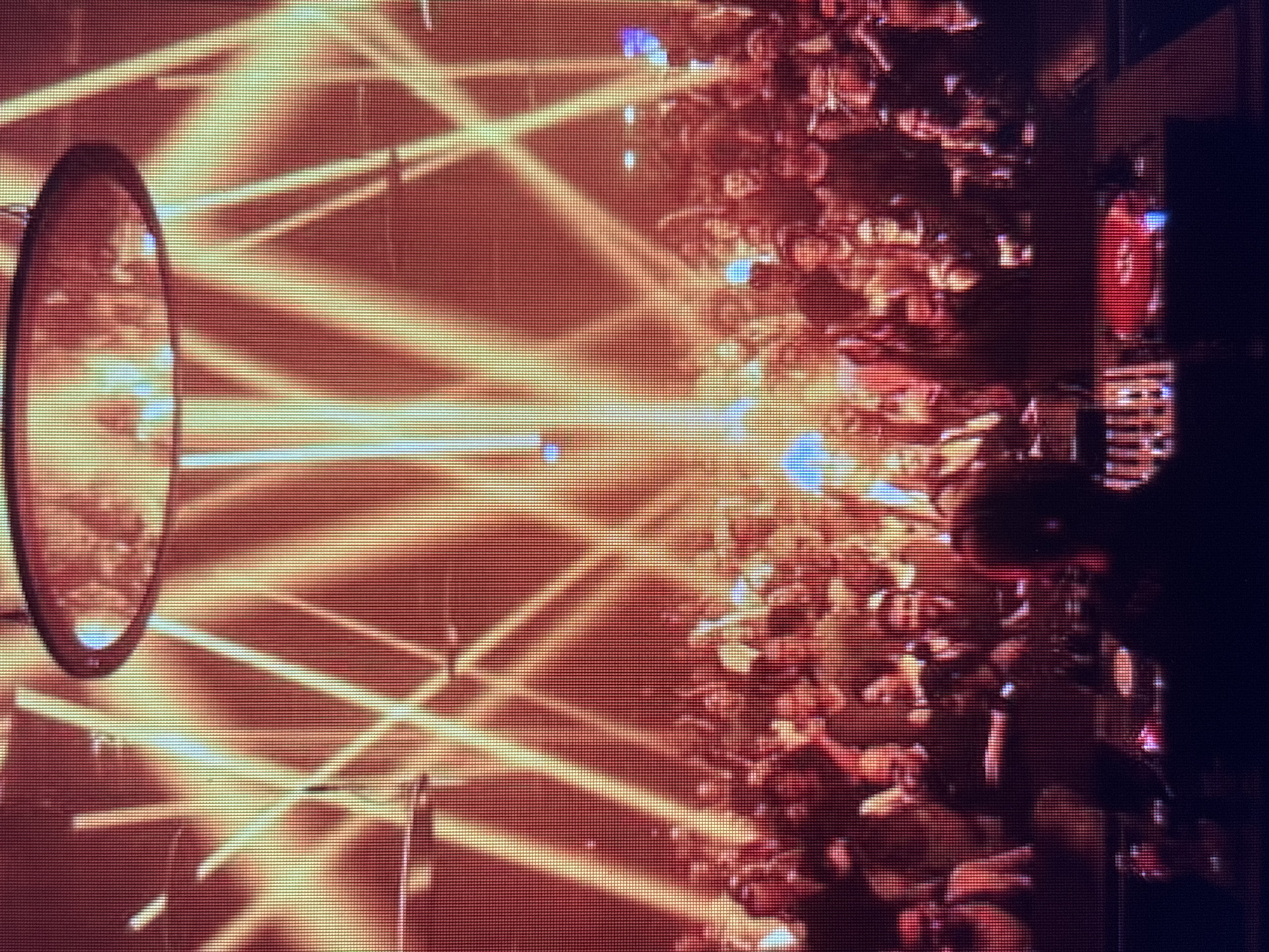 The image depicts a dimly lit indoor venue, possibly a nightclub or concert hall, with a large crowd of people attending an event. The environment is filled with dramatic lighting effects, primarily bright orange and yellow beams emanating from above and crossing each other in a criss-cross pattern. The beams illuminate the scene from multiple angles, creating a vibrant and energetic atmosphere.
In the foreground, silhouetted figures are visible, suggesting that the photograph was taken from within the crowd or from a raised vantage point. One prominent figure, also in silhouette, appears to be standing near what might be a DJ booth or a stage, with equipment in front of them. The crowd is densely packed, with many individuals raising their arms, indicating excitement or engagement with the performance. Some individuals are clearly visible within the crowd, though their features are obscured by the lighting and distance.
The overall impression is of a lively gathering, characterized by dynamic light displays and a spirited audience. The location is described as being in "Unknown, Unknown," indicating that specific geographical details are not available. The image's resolution suggests it might be a digital capture, possibly from a screen or a lower-quality camera, with a visible pixelated texture.