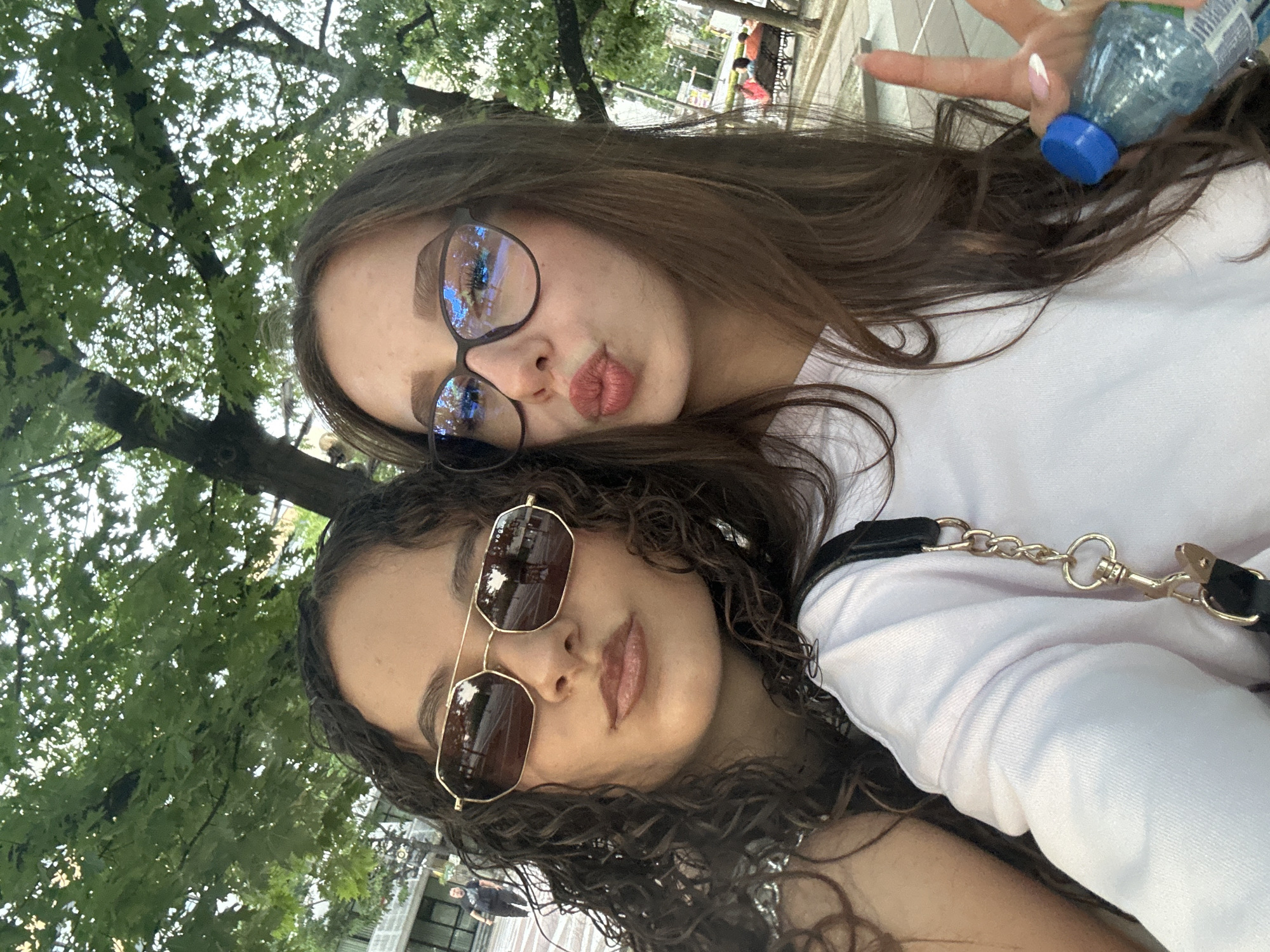 The image captures two young adult females in a close-up outdoor selfie within a public urban environment in Blagoevgrad, Bulgaria. The background features a dense canopy of green deciduous trees, suggesting a park or tree-lined street, with diffuse daylight.
The female on the left possesses dark, curly hair and wears gold-framed hexagonal sunglasses with dark lenses, bearing the "Vogue" brand on the visible right lens. Her lips are slightly parted and appear to have light brown lipstick. She wears a light-colored, long-sleeved garment, and a black strap with a gold chain and clasp, likely from a bag, is visible over her right shoulder.
The female on the right has long, straight brown hair and wears dark-framed spectacles, with prominent blue light reflections on the lenses. Her lips are puckered in a "kissing" pose. Her right hand, featuring manicured nails with a white tip design, holds a small, clear plastic water bottle with a blue screw cap, while simultaneously forming a "peace" sign with the index and middle fingers extended. She also wears a light-colored, long-sleeved garment.
In the background, behind the left subject, a paved area is visible with a male individual in a dark shirt walking in the distance, adjacent to a light-colored building. To the far right of the frame, a dark park bench with two or three seated individuals is discernible on another paved surface amidst additional trees.