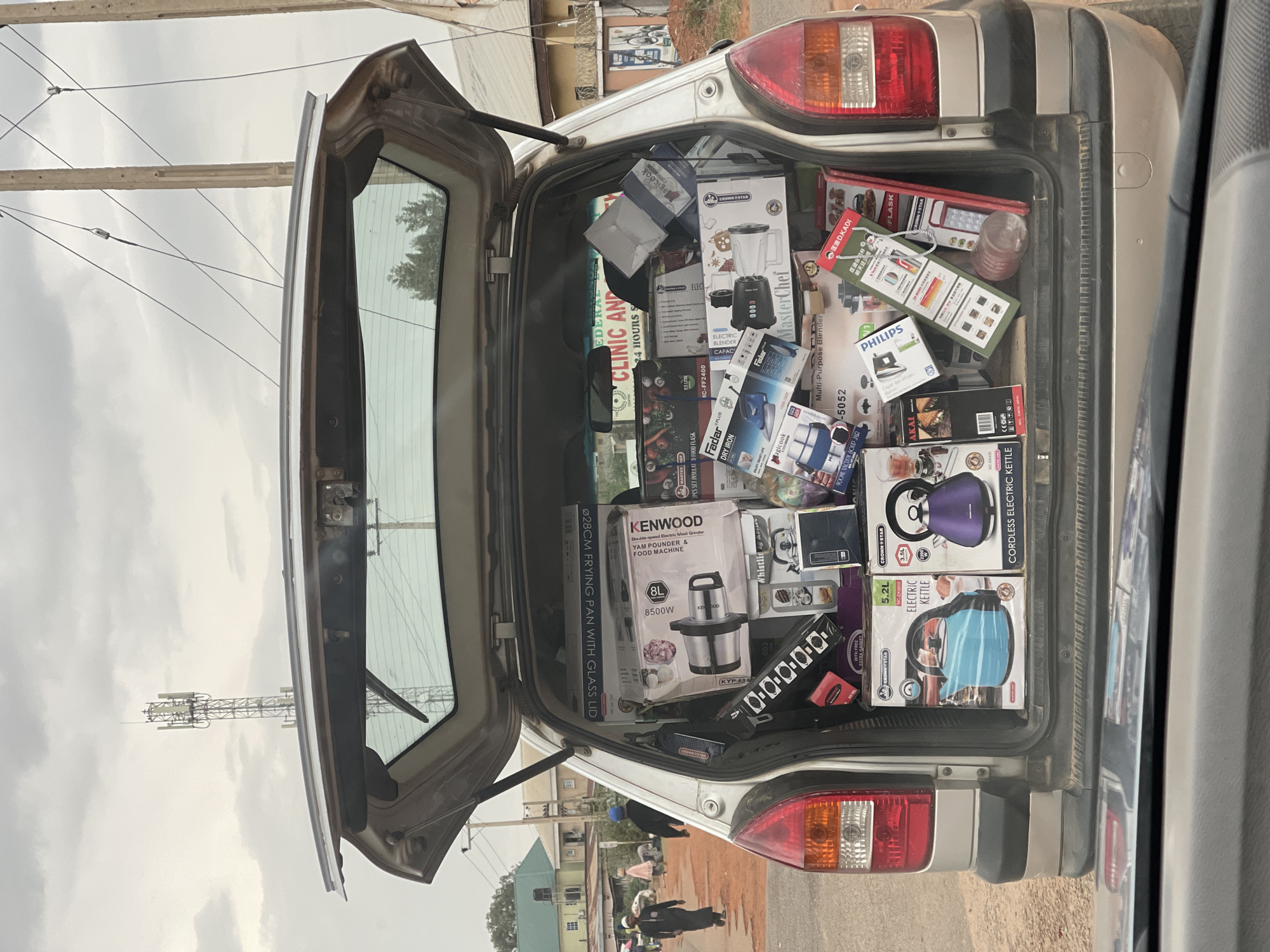 The image shows the open trunk of a silver car filled with boxes of small kitchen appliances. Brands such as Kenwood, Philips, Fadar, and Crown Star are visible. The street scene outside the trunk shows a road, buildings, utility poles, and pedestrians. A sign for the "Federal Clinic and" is partially visible. The location appears to be Jos, Nigeria, based on the visible signage and the products, clothing, and infrastructure.