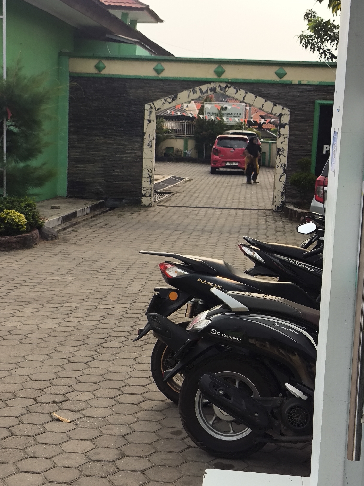 The image shows an exterior view of the Cilegon City Haj Information Center, located in Cilegon, Indonesia. The foreground features several parked black motorcycles, including models labeled "NMAX," "VARIO," and "SCOOPY." The motorcycles are parked on a paved courtyard made of hexagonal interlocking pavers. In the background, an arched gateway is visible, decorated with green and yellow accents and dark stone cladding. Beyond the arch, a red car is parked, and two individuals are walking away from it. A sign above the arch reads "PUSAT INFORMASI HAJI KANTOR KEMENTERIAN AGAMA." The scene is lit by natural daylight, suggesting it is daytime. The overall environment appears to be an institutional building's entrance or courtyard.