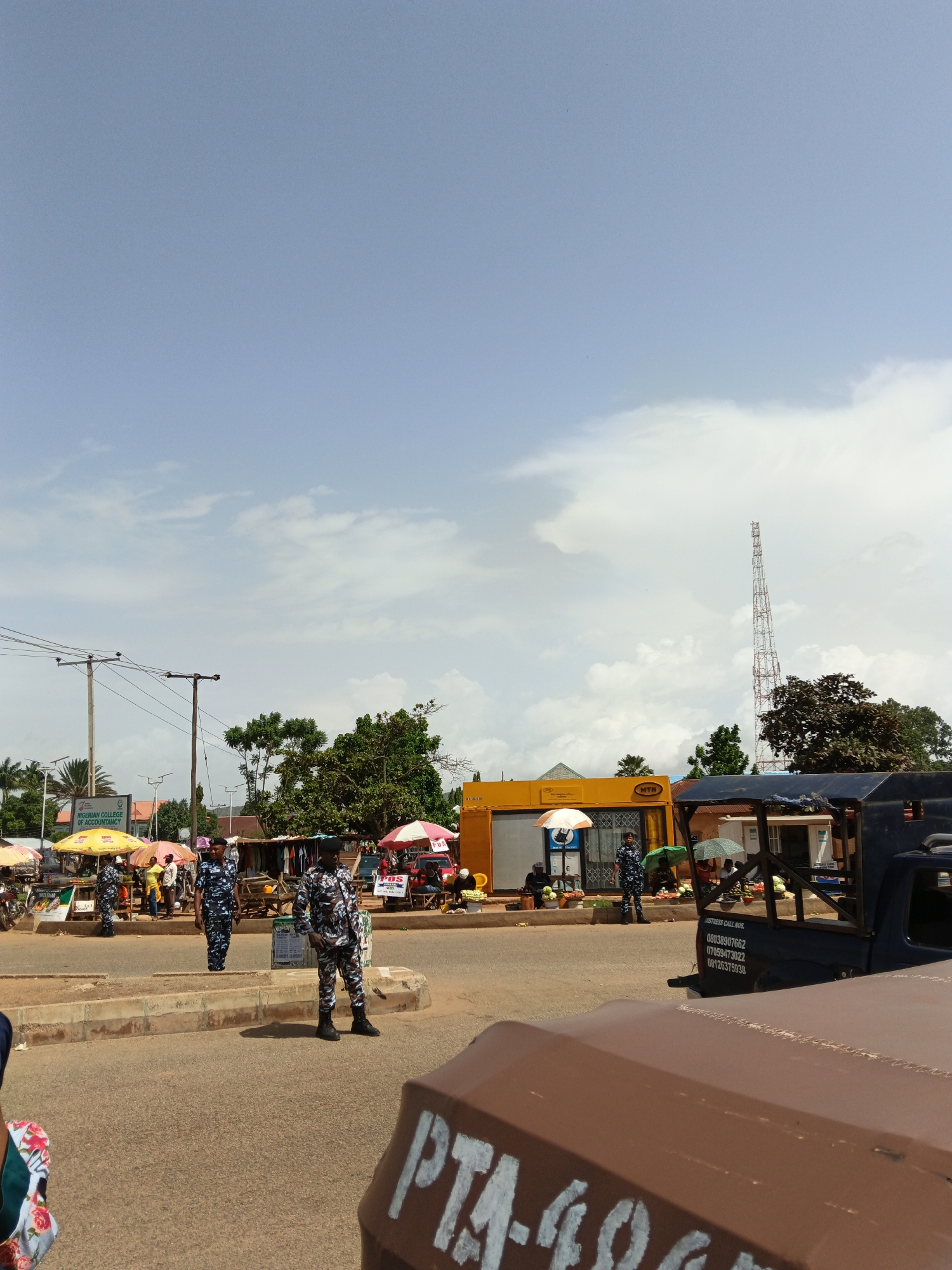 The image captures an outdoor scene in what appears to be a market or roadside area in a city. The sky is partly cloudy, with a tall communication tower visible in the distance. In the foreground, a brown vehicle is partially visible, with markings "PTA-499" painted on its surface.
Several uniformed individuals, likely law enforcement or security personnel, are present. One individual stands on the side of the road, while another is positioned further back. They are wearing camouflage-patterned uniforms and dark boots.
The middle ground of the image features market stalls with umbrellas providing shade. Various items are displayed for sale, including fruits and clothing. A yellow building with "MTN" signage is also visible, possibly a business establishment. A blue truck is parked near the market stalls, with contact numbers listed on its rear.
The overall scene suggests a bustling commercial area, possibly with a security presence to maintain order. The environment is characterized by paved roads, market infrastructure, and urban foliage.