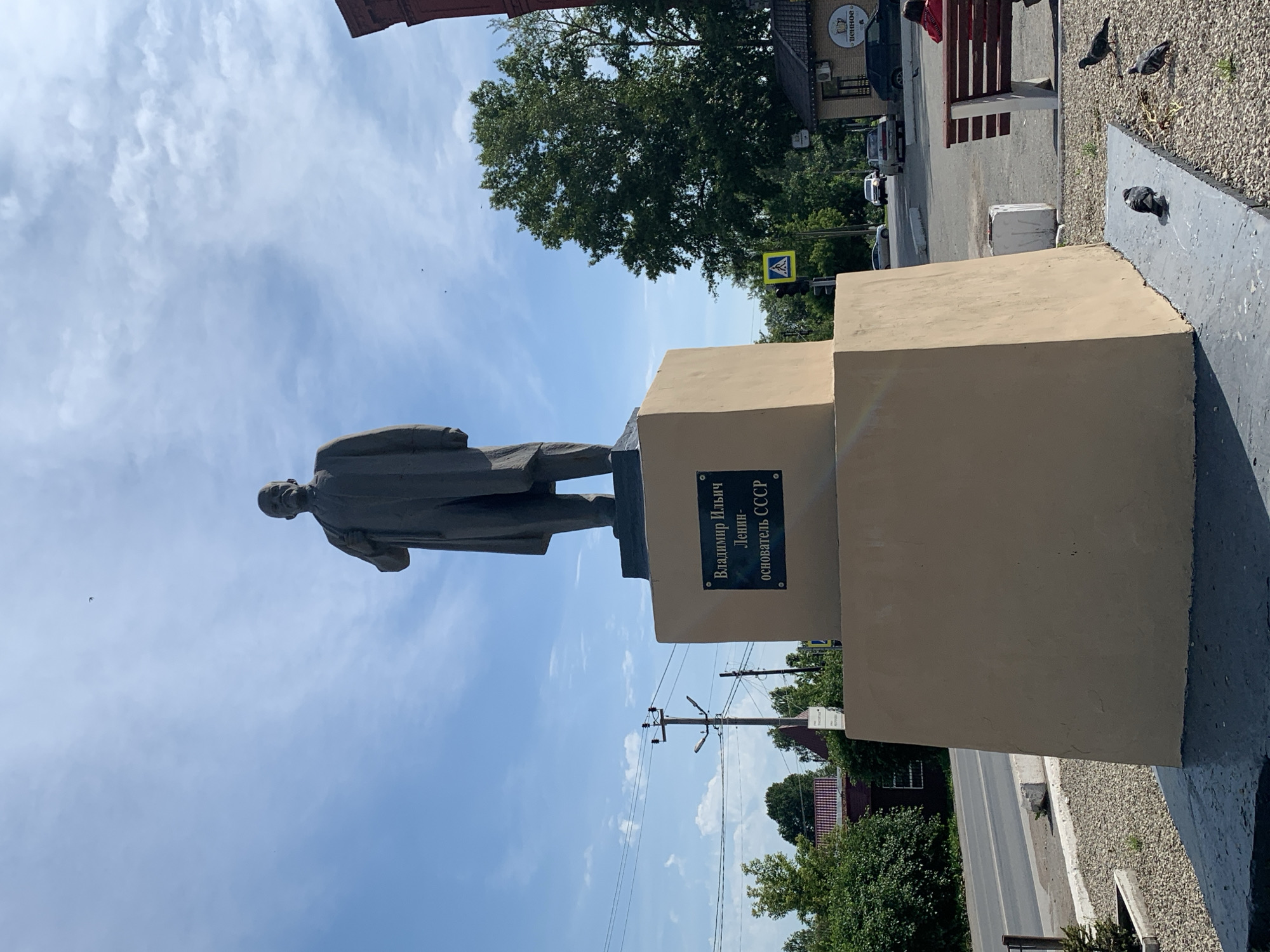 An outdoor, eye-level, medium shot captures a statue of Vladimir Lenin in Kungur, Russia. The monument, made of dark grey material, depicts Lenin standing, with his right arm raised and his left hand on his chest. He wears a suit and a stern expression. The statue stands on a two-tiered, beige pedestal. The lower tier is larger and more substantial, while the upper tier is smaller and rectangular. A dark plaque with Russian text is affixed to the front of the upper tier. The text reads: "Владимир Ильич Ленин - основатель СССР" which translates to "Vladimir Ilyich Lenin - Founder of the USSR."
The statue is situated in an open area with a gravel ground surface. A paved pathway runs along the base of the pedestal. In the background, a street with passing cars is visible, lined by trees and buildings. A red-brick building is partially visible to the right of the statue. A wooden bench with a person sitting on it is to the right, and several pigeons are on the ground near the statue. The sky is blue with scattered clouds, and sunlight illuminates the scene. A traffic light and a pedestrian crossing sign are visible on the street.