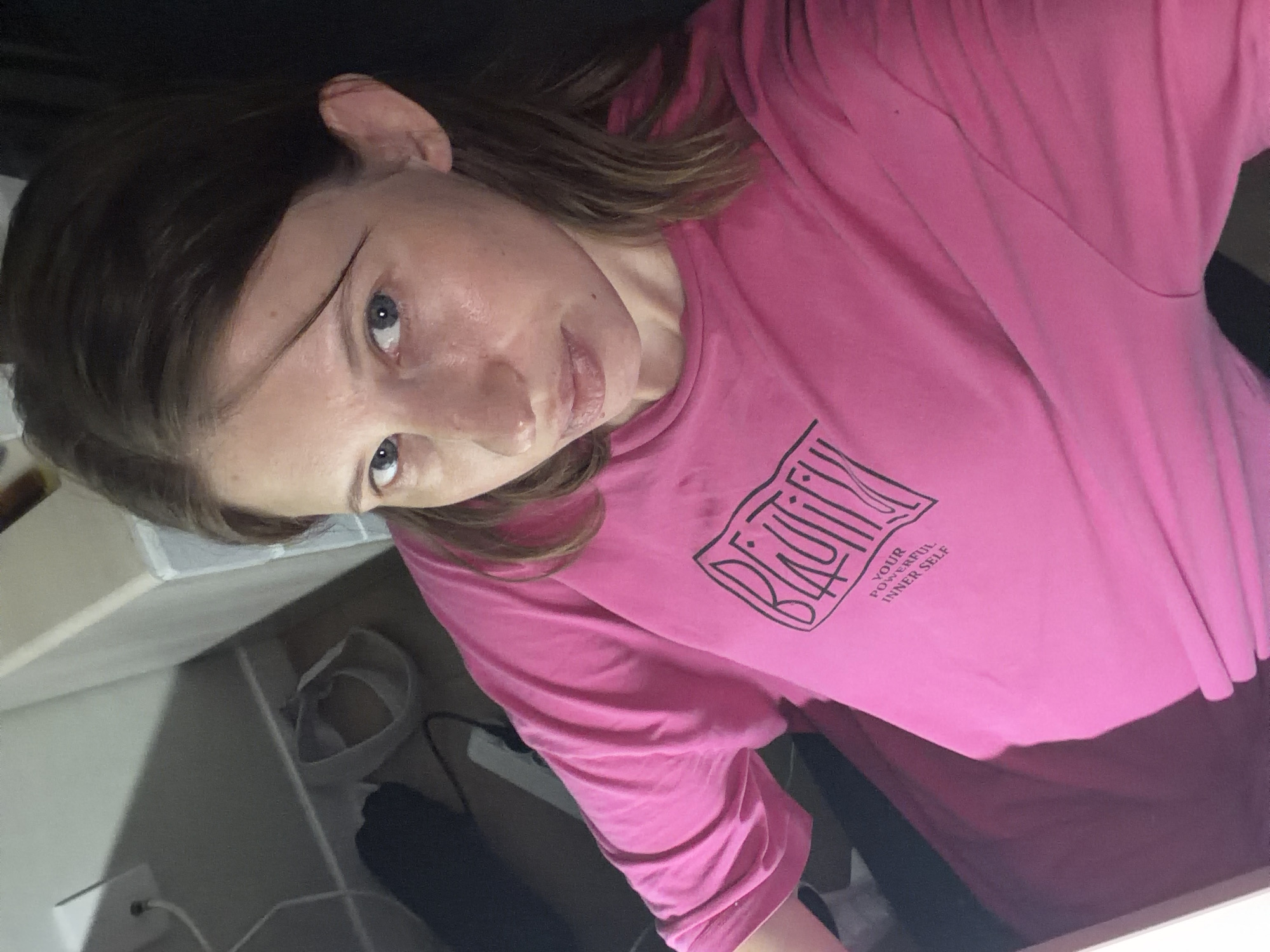 A young woman with brown hair and blue eyes is wearing a bright pink t-shirt with a graphic design. The t-shirt has the word "BEAUTIFUL" in large, stylized lettering, with smaller text below it that reads "YOUR POWERFUL INNER SELF." The woman is looking at the camera with a neutral expression. She appears to be indoors, possibly in a bedroom or living area, with a white dresser or shelf in the background and a pair of gray sandals on the floor. The lighting is dim, and the image is taken from a slightly high angle.