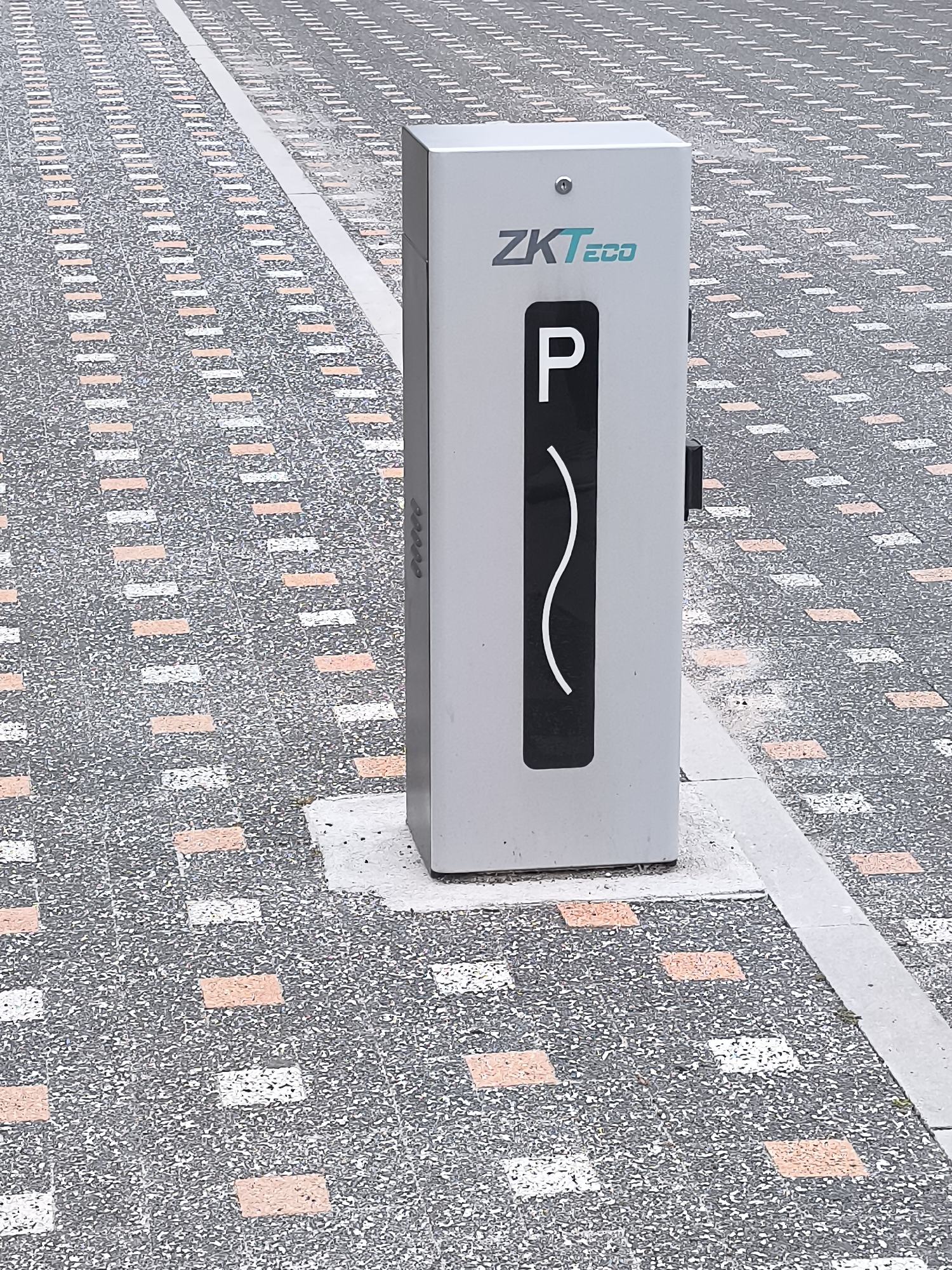 The image shows a vertical, light-colored parking payment terminal with the "ZKTECO" logo in teal lettering and a black panel with a white wavy line and a white "P" inside it. The terminal is situated on a paved surface composed of dark gray aggregate interspersed with regularly spaced rows of small, rectangular tiles in peach and white. A light gray concrete strip runs diagonally across the paved area, separating it from another section of the same paving pattern. The scene is outdoors, with the paving extending into the background, suggesting a public area such as a sidewalk, plaza, or parking lot. The light appears to be diffused, possibly indicating an overcast day. The terminal is firmly planted in the ground, anchored by a concrete base. There are no people visible in the image, and no actions are taking place. The environment suggests an urban setting within the city of Bălți, Moldova, characterized by its distinctive paving style.