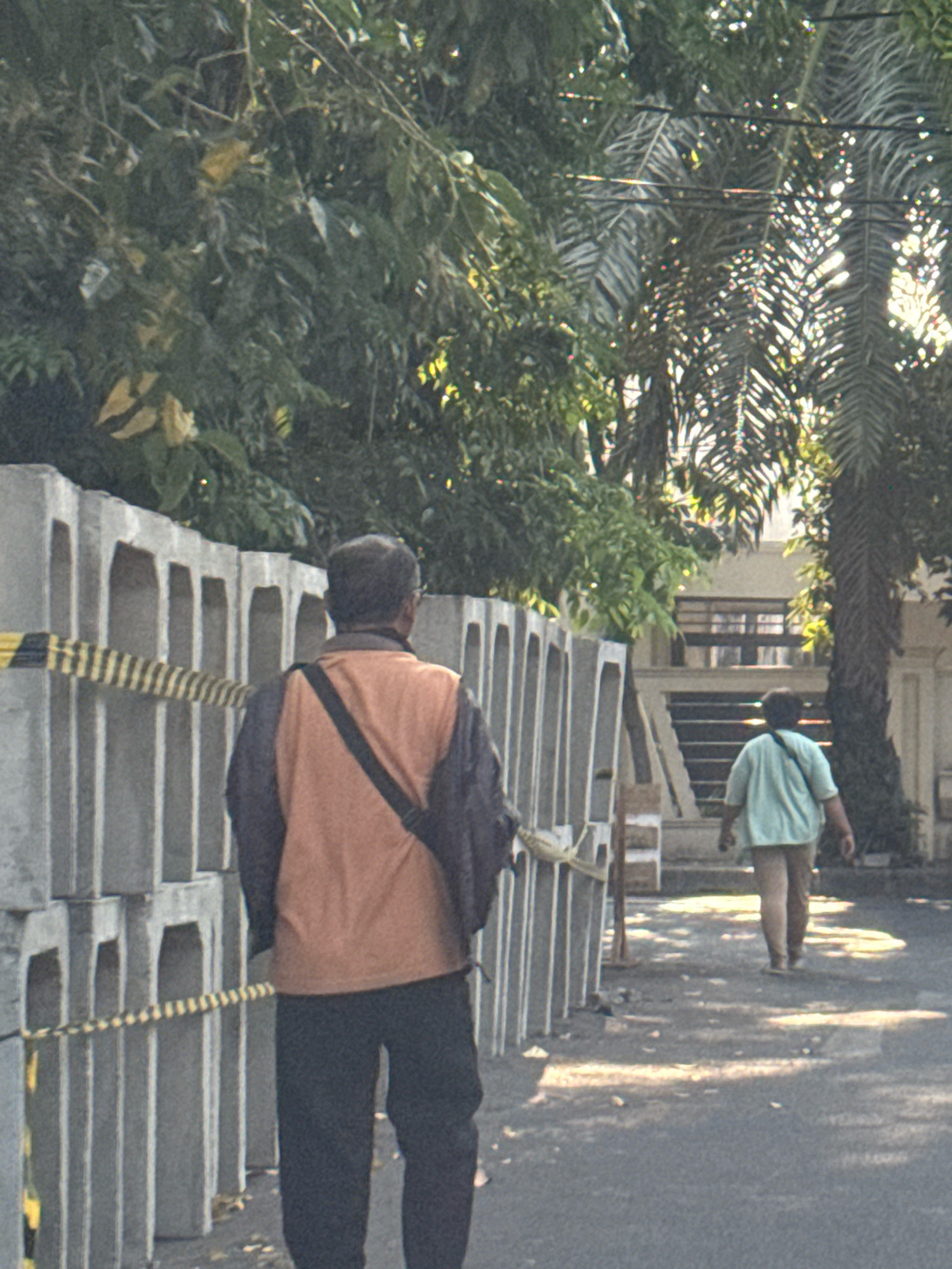 The image depicts an outdoor scene on a sunny day in Surabaya, Indonesia. In the foreground, a man wearing a dark jacket over an orange vest and black pants walks away from the camera. He has a dark bag slung across his shoulder. To his left, a row of stacked concrete barriers is visible, with a yellow and black striped caution tape stretched across them. In the background, another person, seen from behind, walks away from the camera wearing a light blue t-shirt and light brown pants. This person also has a dark bag slung across their shoulder. Behind the second person, there appears to be a building with stairs leading up to it. Lush green foliage and palm trees are scattered throughout the scene, with sunlight casting dappled shadows on the asphalt road. The overall impression is of a construction or maintenance area with people walking through it.