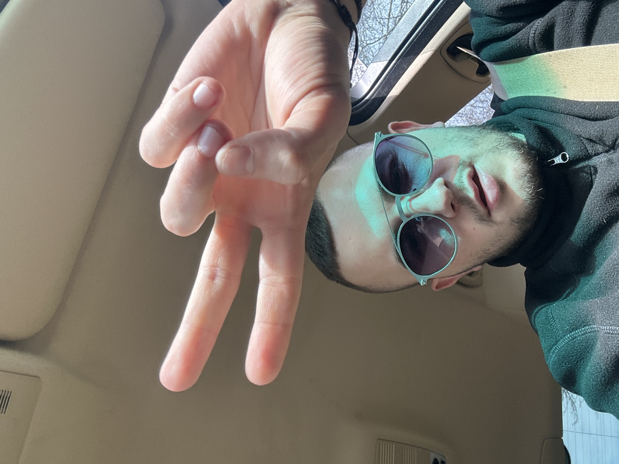 A man is taking a selfie from inside a car. He is wearing round sunglasses with silver frames and a black hoodie. His right hand is raised, forming a gesture where the index, middle, and thumb fingers are extended while the ring and pinky fingers are folded into the palm. He is wearing a wrist band on the right arm. The car interior is beige, and a seatbelt is visible across the chest. The photograph appears to be taken looking up into the roof of the vehicle, the car is likely in Plovdiv, Bulgaria.