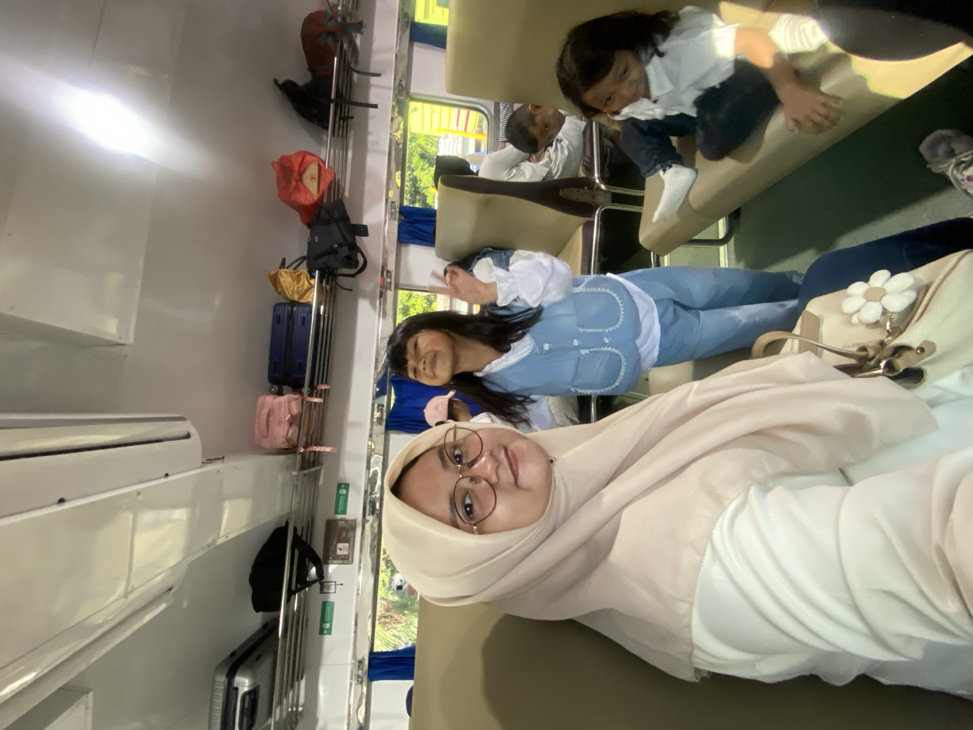 The image shows a group of people in what appears to be a train carriage. The foreground is dominated by a person wearing a hijab and glasses. Behind them, another person is giving a peace sign. In the background, there are two other people, one reclining on the seat and the other partially visible. Luggage is visible on the overhead rack. The setting seems to be in Probolinggo, Indonesia, based on the train carriage interior.