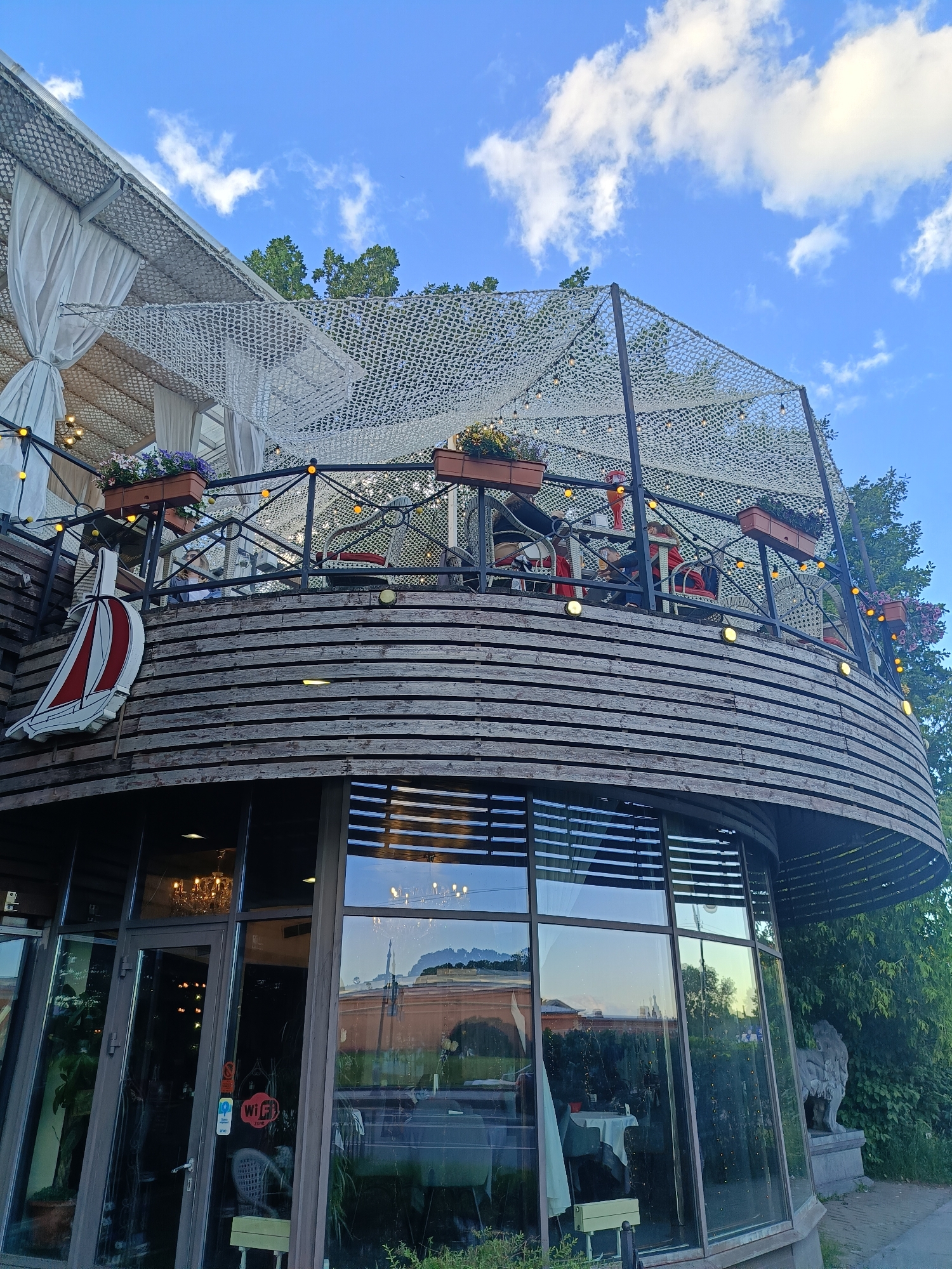 This media file captures a picturesque exterior view of a waterfront restaurant, likely situated in Saint Petersburg, Russia. The building features a distinctive curved, multi-story design with a wooden exterior, reminiscent of a ship's hull. A second-story balcony is visible, adorned with potted plants, string lights, and a white, netted canopy overhead. The balcony railing is made of dark metal, and several tables and chairs are arranged on the deck, suggesting outdoor seating. A prominent boat emblem with red and white sails is affixed to the side of the building.
The ground floor is characterized by large, floor-to-ceiling windows, reflecting the surrounding greenery and sky. Through the glass, glimpses of the restaurant's interior can be seen, including chandeliers and tables set with white tablecloths. A statue of a lion stands guard near the corner of the building. Visible text includes "Wi-Fi ZONE" stickers on the glass door.
The time of day appears to be late afternoon or early evening, with the golden light of the setting sun casting a warm glow and creating reflections in the windows. The sky is a vibrant blue, dotted with fluffy white clouds, indicating pleasant weather. The lush green trees surrounding the establishment add to the serene and inviting atmosphere of the location. The overall impression is one of a charming and established dining establishment by the water, possibly a popular spot for patrons to enjoy the view and ambiance.
