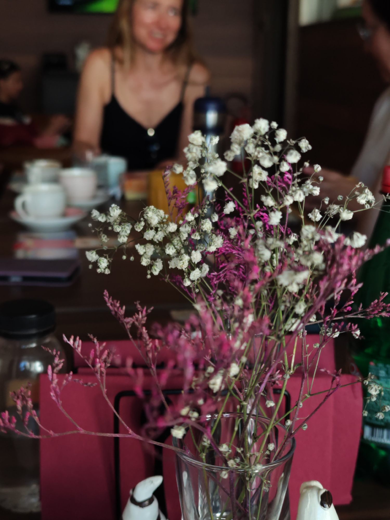 In an indoor setting, possibly a cafe or restaurant in Budva, Montenegro, a woman with blonde hair and glasses smiles while wearing a black tank top. In the foreground, a bouquet of small white and pink flowers is arranged in a glass vase. On the table, several white teacups and saucers are visible, along with a dark-colored bottle and a dark-colored object that could be a phone. A red napkin is placed behind the flowers.