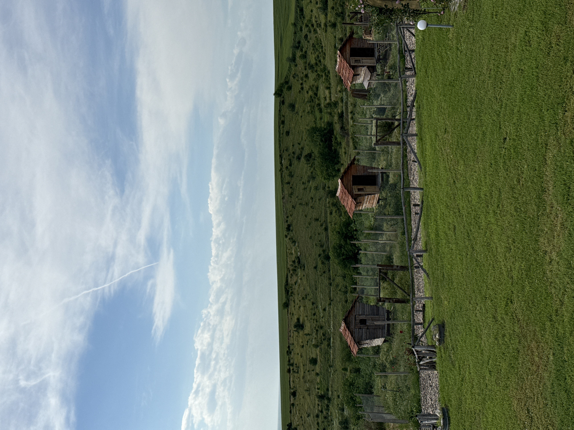 The image shows a landscape scene. On the left is a blue sky with white clouds, including a contrail. The right side depicts a grassy field with three small, wooden structures, possibly sheds or small houses, aligned in a row. They have reddish-brown roofs and are enclosed by wooden fences with wire mesh. A path or stone walkway runs alongside the sheds. Lush green grass covers the immediate foreground. The location is in Mechka, Bulgaria.