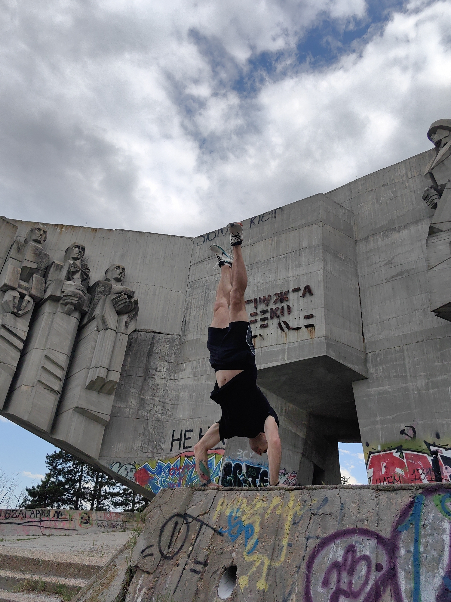 A man is performing a handstand on a graffitied concrete structure in Varna, Bulgaria. The structure is part of a large concrete monument with Soviet-era style figures carved into its surface. The monument also has graffiti and writing on it. The sky is overcast with clouds.