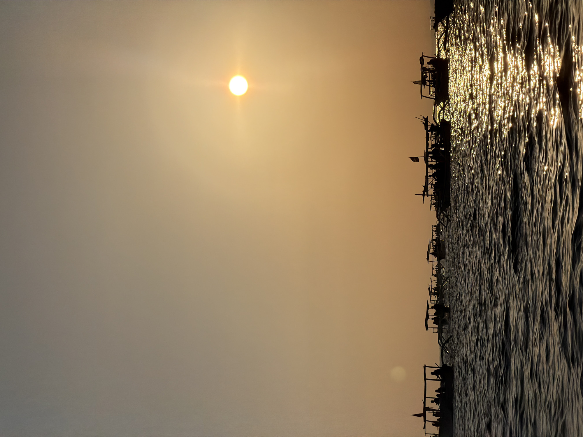 At sunset in Buleleng, Indonesia, several silhouetted boats are moored on the ocean. The sun, a bright orb with rays, is low in the orange and yellow sky. The water reflects the sun's light, creating a shimmering effect. People are visible as dark figures on the decks of the boats, some appearing to be engaged in conversations or activities. The boats themselves are long and narrow, with awnings providing shade.