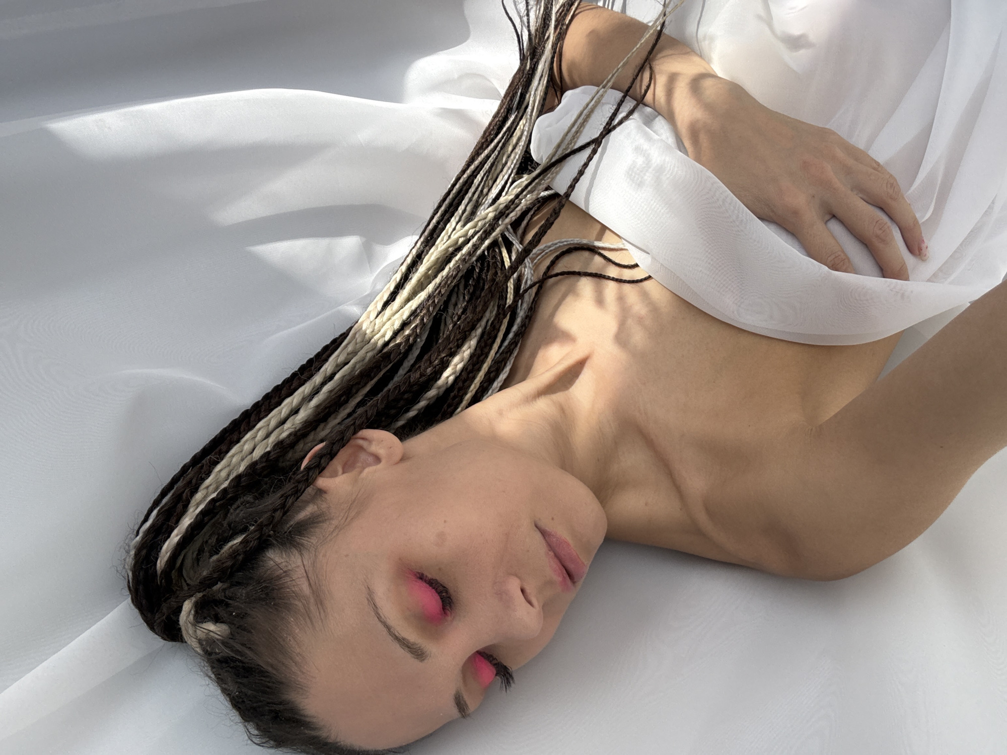 A woman with dark and blonde braided hair is lying on a white fabric surface. She has pink makeup on her eyelids. She is holding the white fabric. The location of the photograph is unknown.