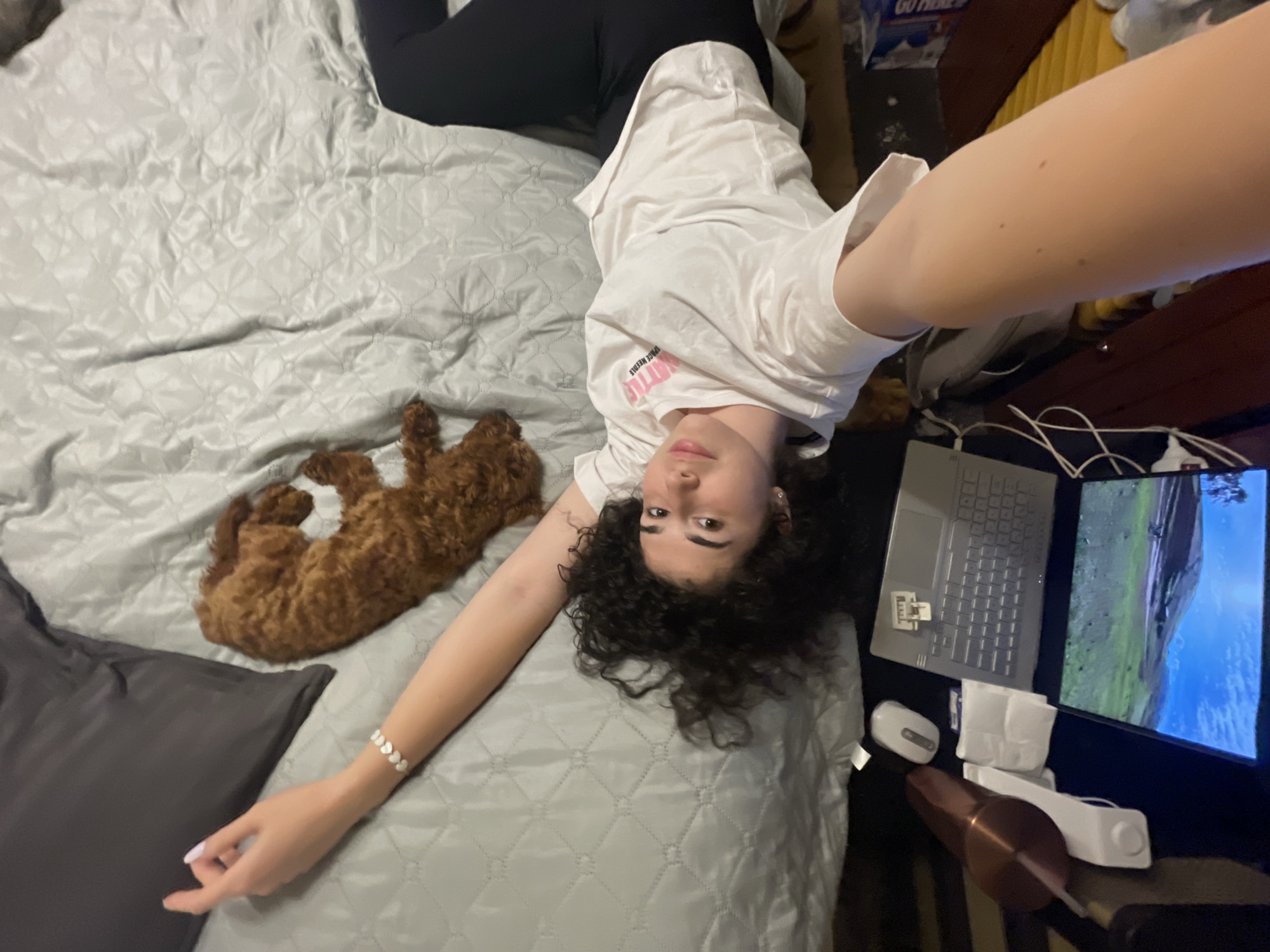 A woman with curly hair is lying on a bed with a light gray quilted bedspread, taking a selfie. She is wearing a white t-shirt and black pants. Next to her is a small, brown, poodle-like dog curled up on the bed. A gray pillow is visible near her hand. To the right, a laptop and monitor are on a surface next to the bed. The monitor displays a landscape scene with mountains and blue sky. A white mouse, tissues, and a copper-colored tumbler are also visible. The scene does not contain enough information to determine the location in Sofia, Bulgaria.