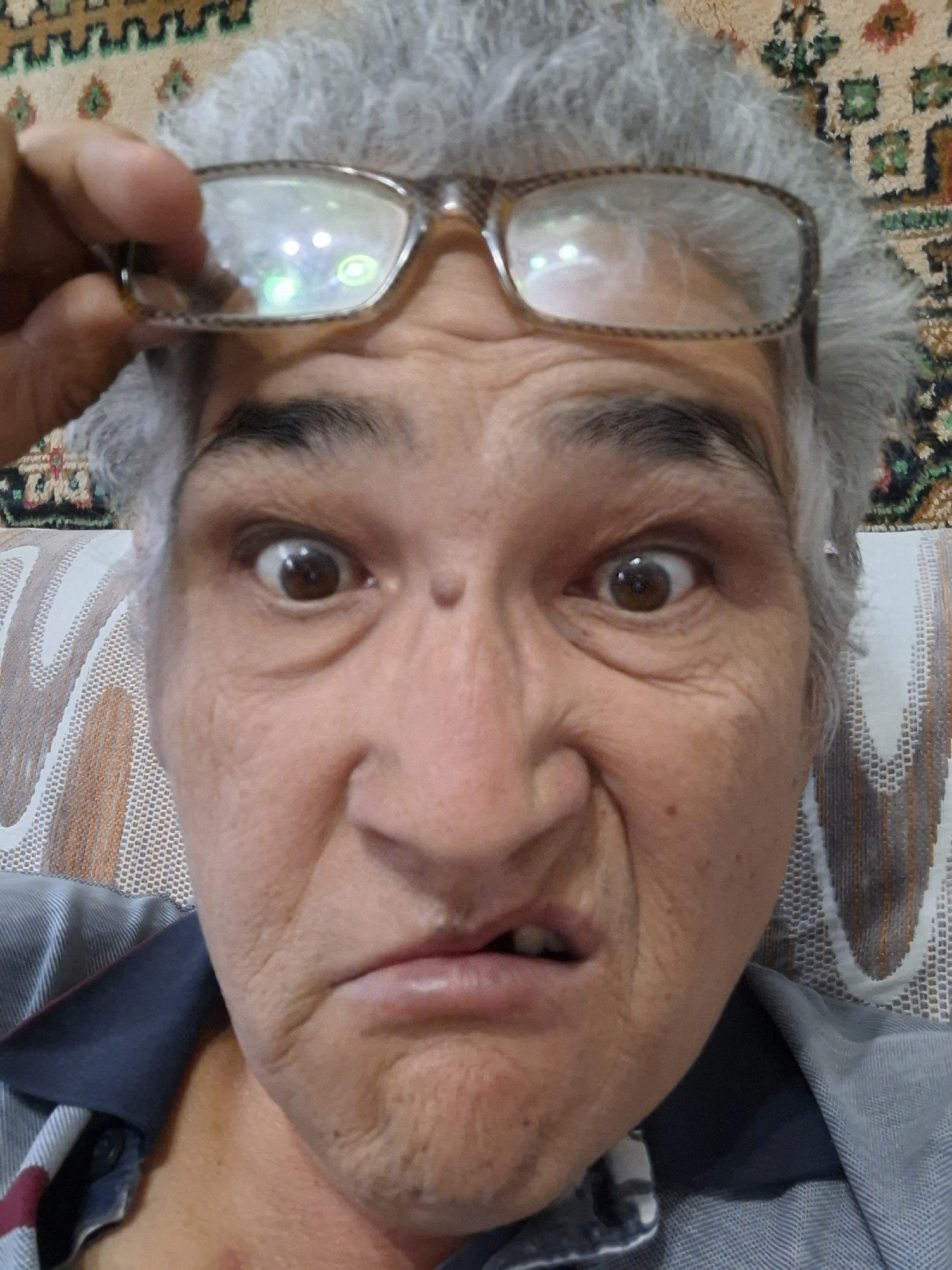 A close-up, eye-level, selfie-style photograph shows a person's face as they make a distorted facial expression. The person, who has graying hair and is wearing glasses that are being held up by their hand, is looking directly at the camera. The background is out of focus, but it appears to be an interior setting with a patterned rug on the wall and a couch or chair. There is no indication that this image was taken in Almaty, Kazakhstan.